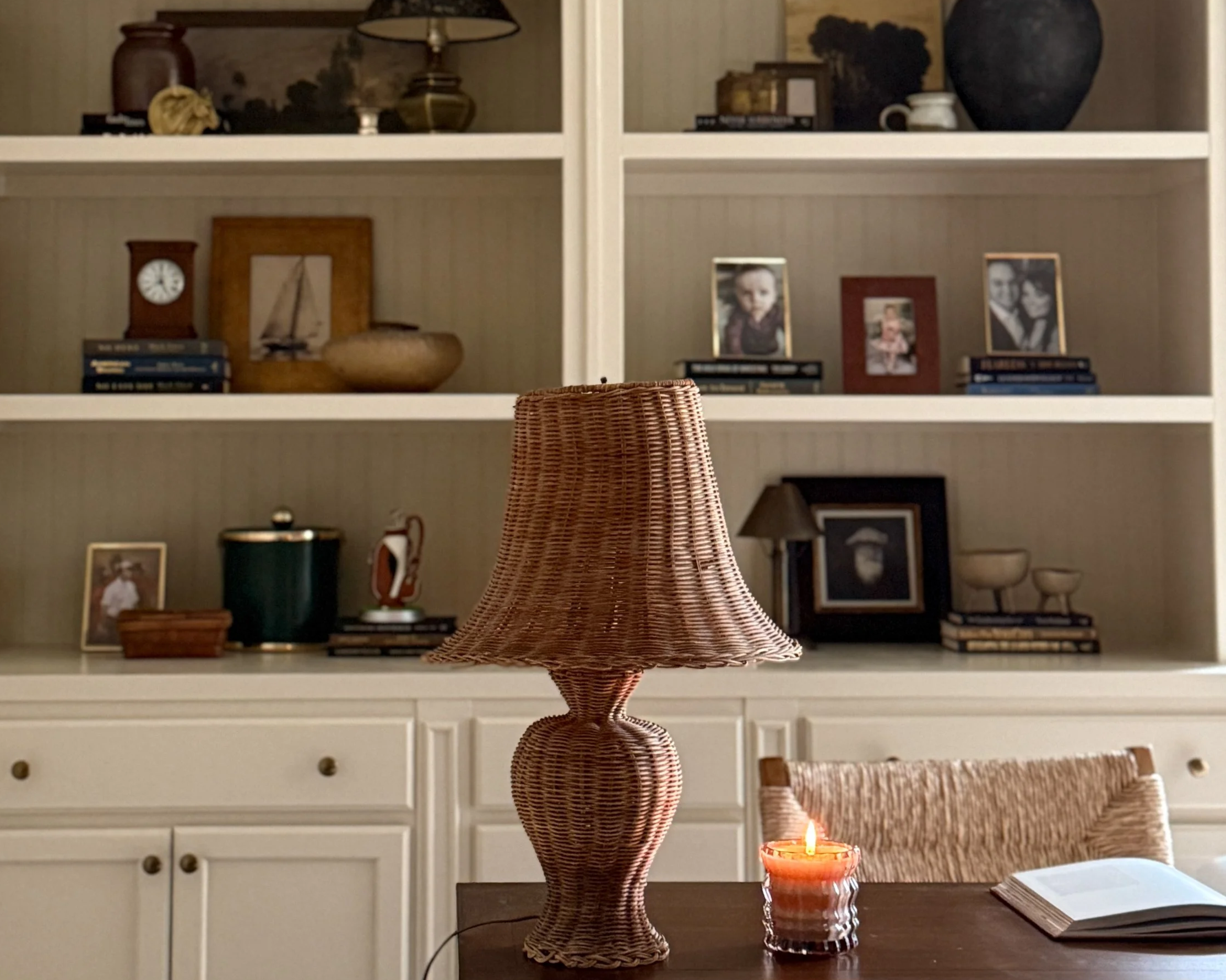 A room with a white bookshelf, decorative objects, framed photos, candles, and a wicker lamp in the foreground.