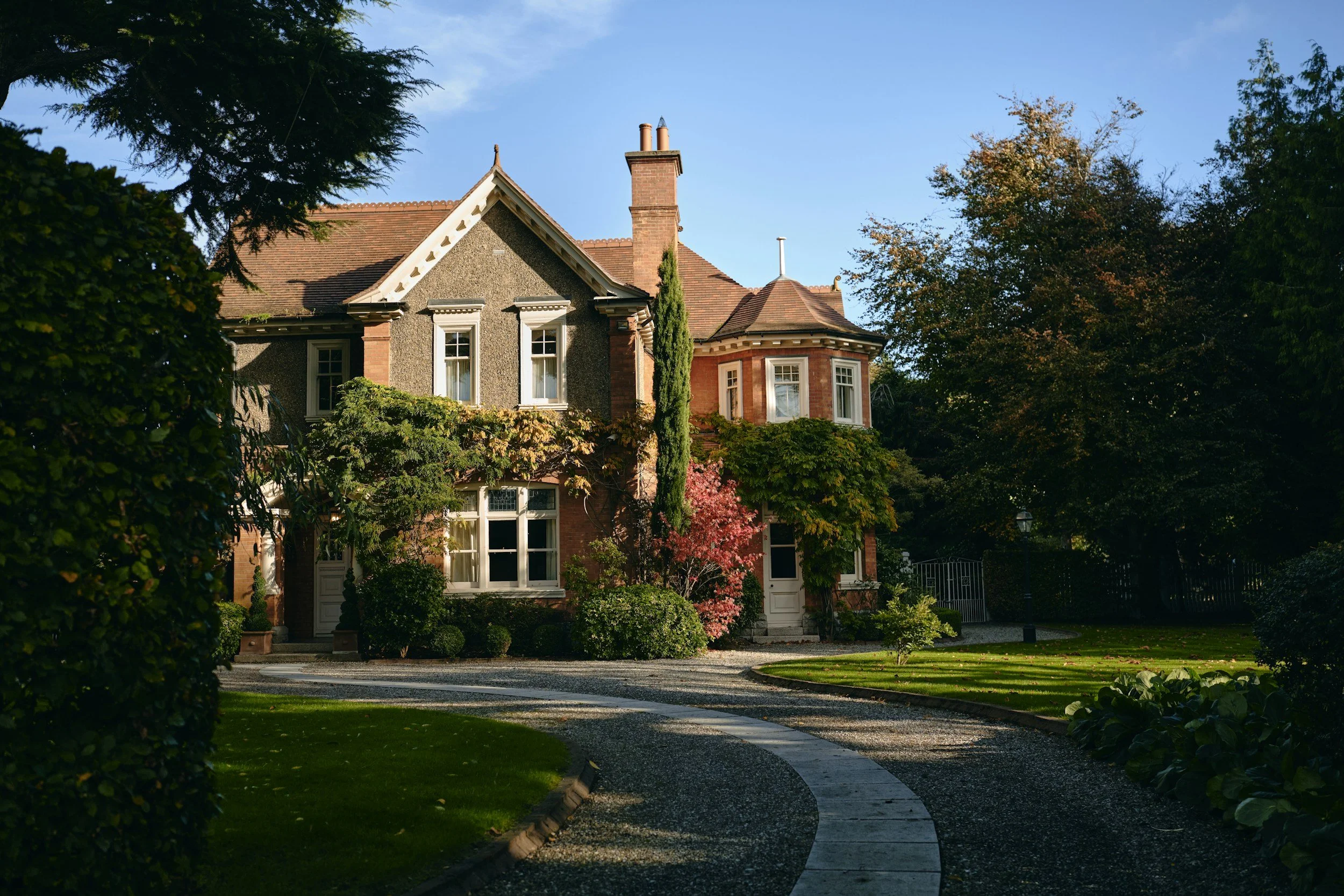 A large house with brick and stucco exterior, surrounded by lush green trees and shrubs, with a curved gravel pathway and a lawn in the front yard, under a clear blue sky.