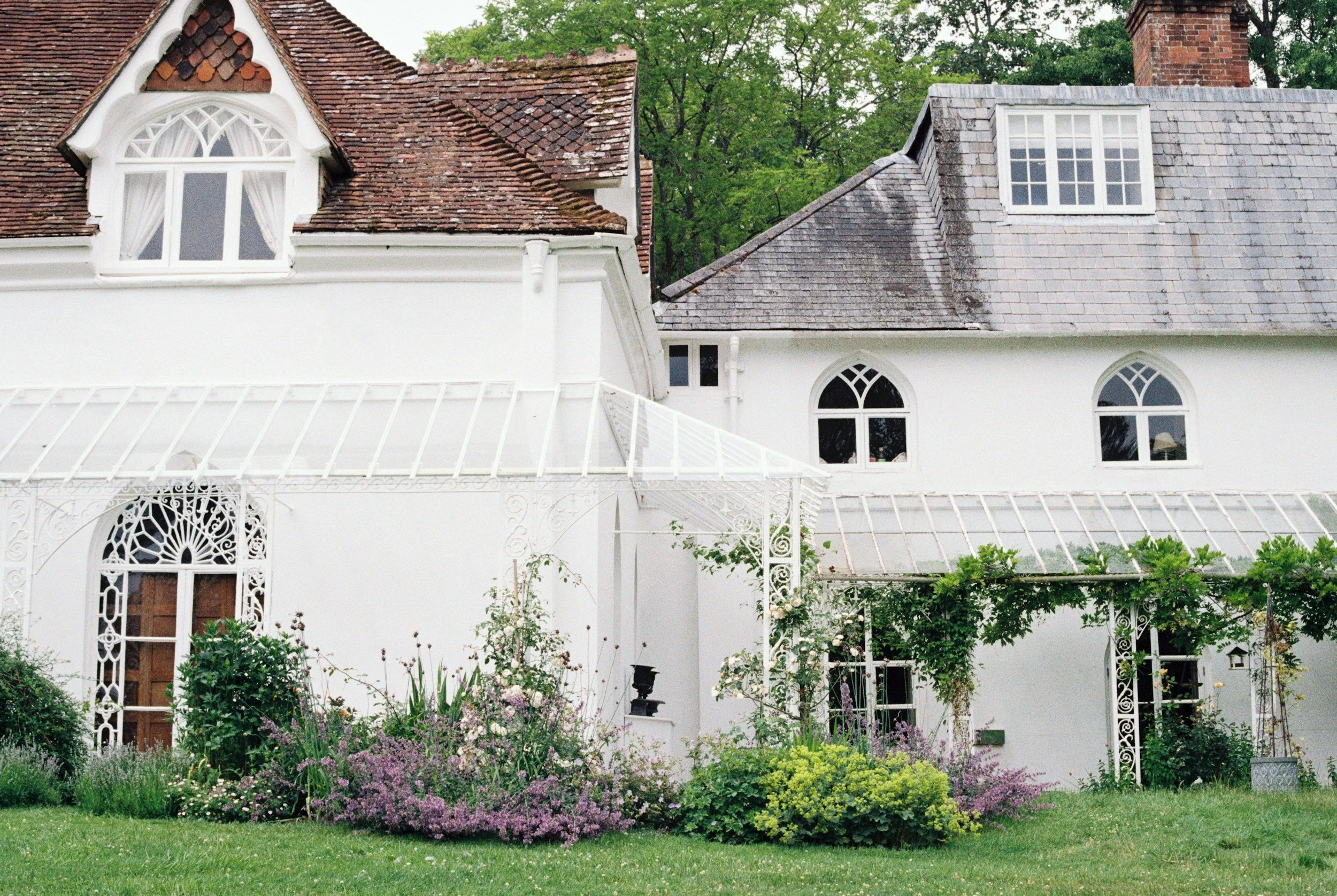 White house with multiple gabled roofs, brick and shingle roofing, decorative ironwork, and flowering garden in the foreground.
