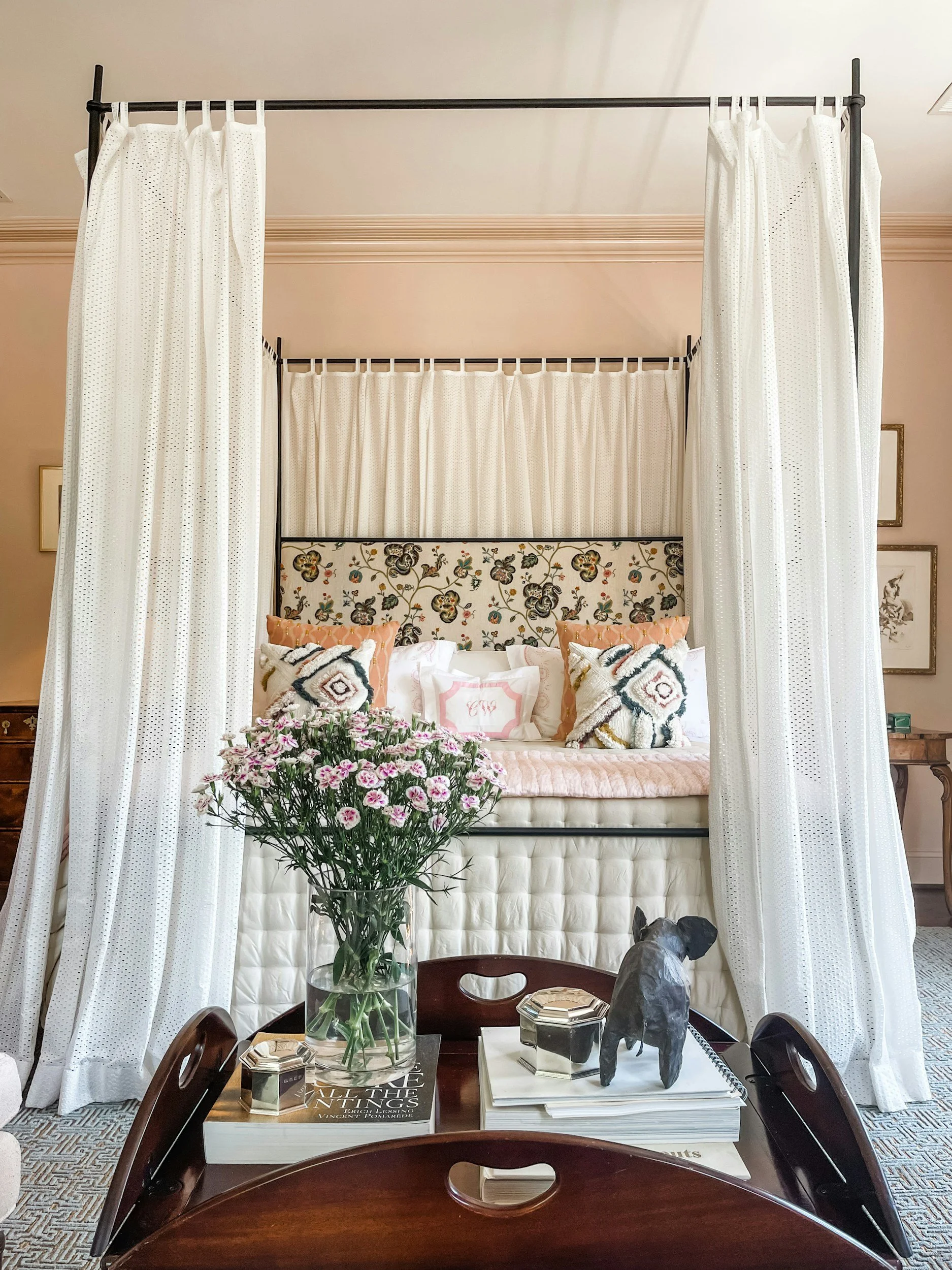 View of a bed with a floral upholstered headboard, surrounded by white curtains. In the foreground, a dark wooden dining table with a glass vase of pink flowers, books, magazines, decorative objects, and a dog sculpture. The room has framed artwork on the walls and a patterned carpet.