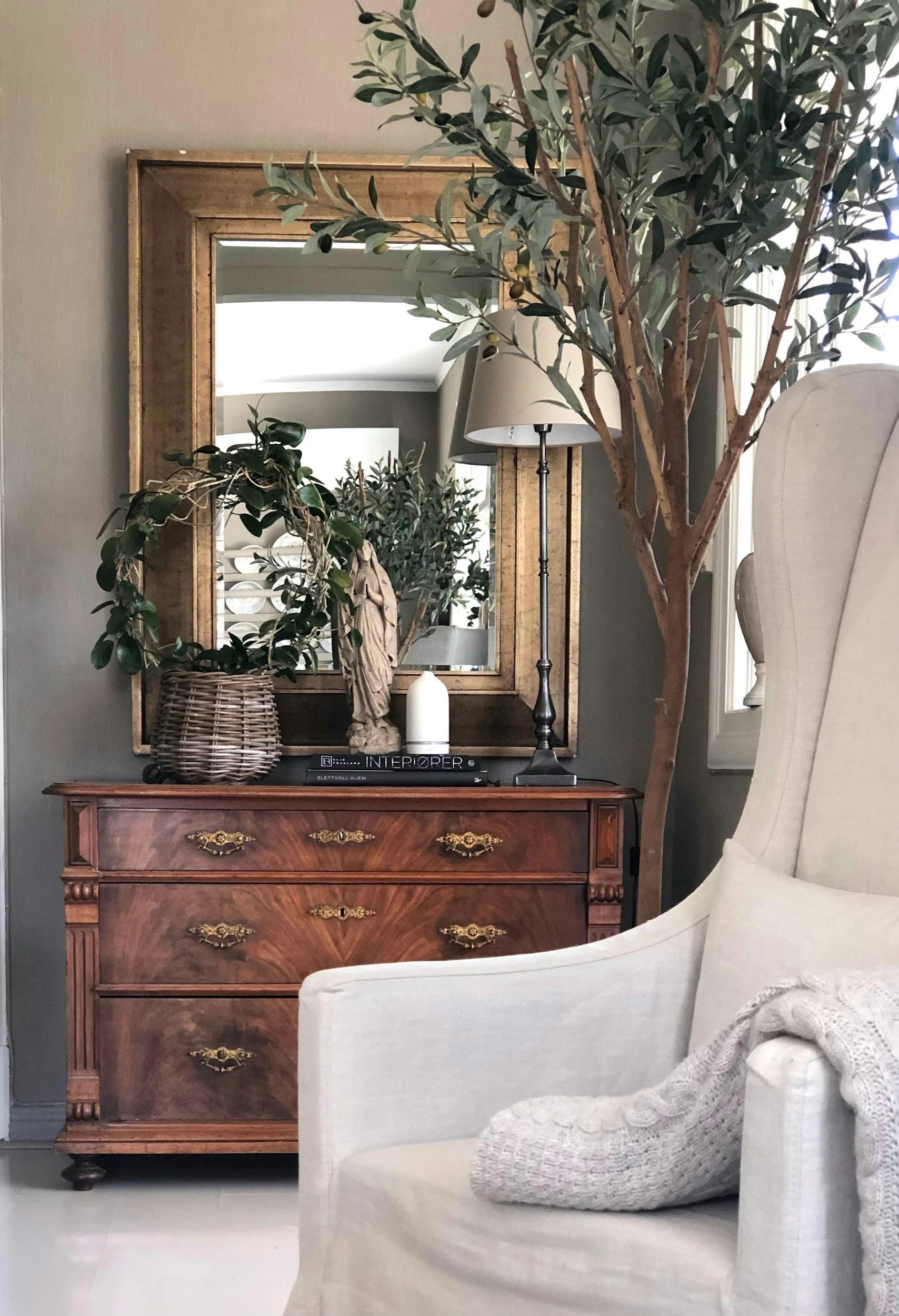 A living room corner with a wooden dresser, a large mirror with a golden frame, a table lamp, a decorative statue, a stack of books, and a potted olive tree. There is also a white upholstered armchair with a knitted blanket and a pillow.