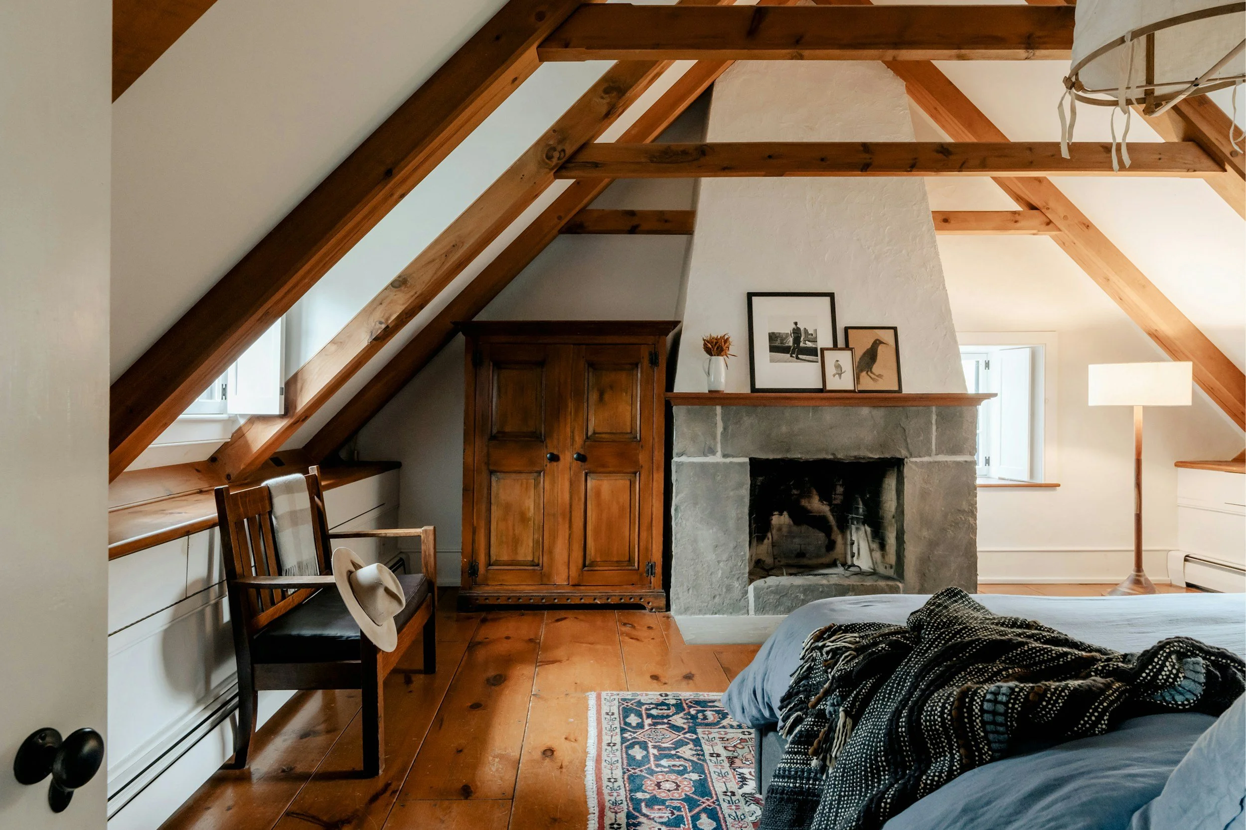 Interior view of a cozy room with slanted wooden ceiling beams, a stone fireplace with framed pictures on the mantel, and a wooden cabinet. There are two windows, a lamp, and a bed with a patterned blanket in the foreground. A small wooden table and mismatched chairs are also visible.