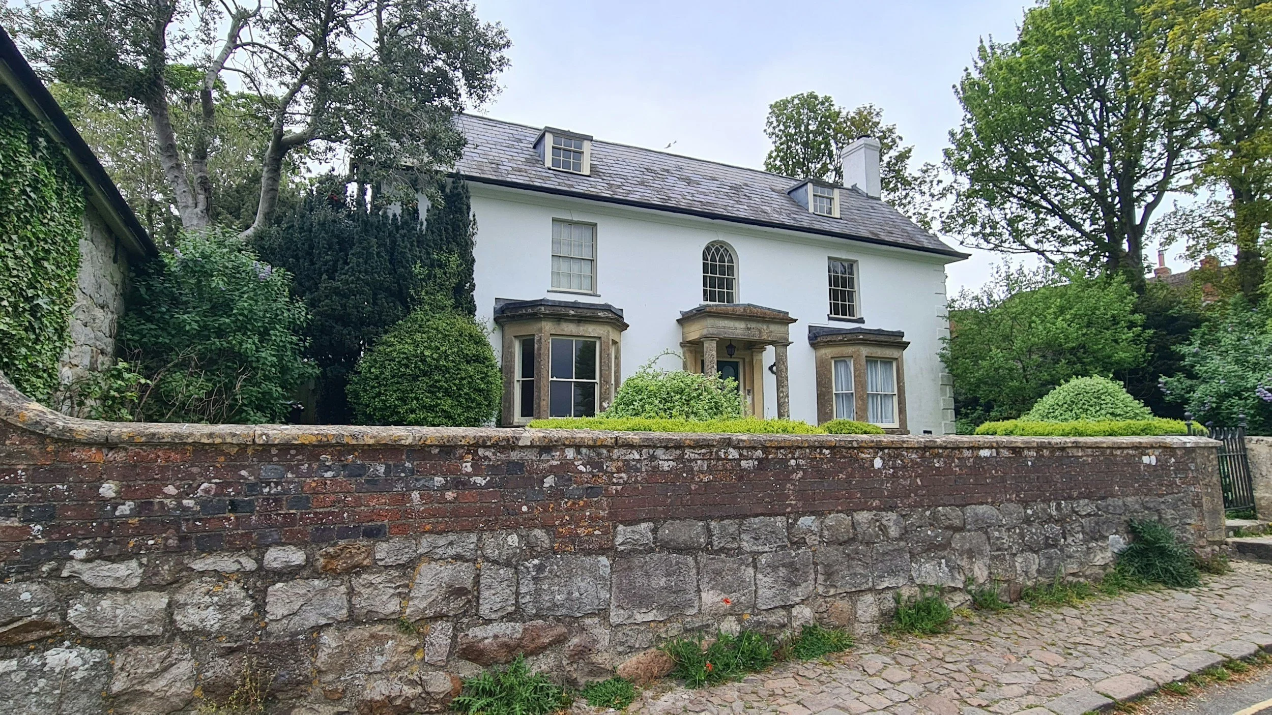 A white two-story house with large windows and a pitched roof, surrounded by trees and greenery, with a brick wall in the foreground and a cobblestone path in front.