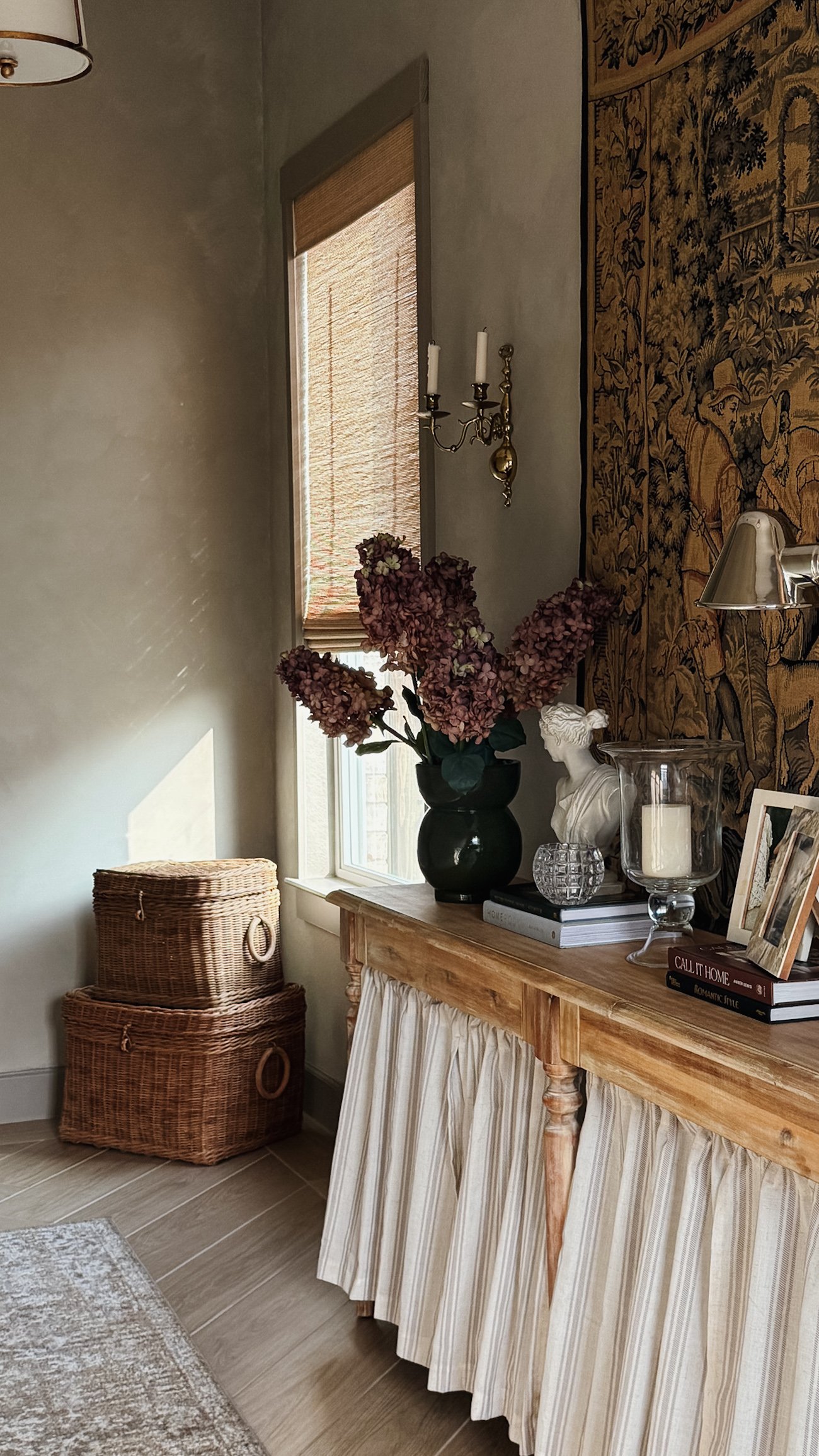 A cozy corner with a wooden sideboard featuring a skirt, a flower arrangement with pink hydrangeas, candles, and books, beside a window and decorative wicker baskets.