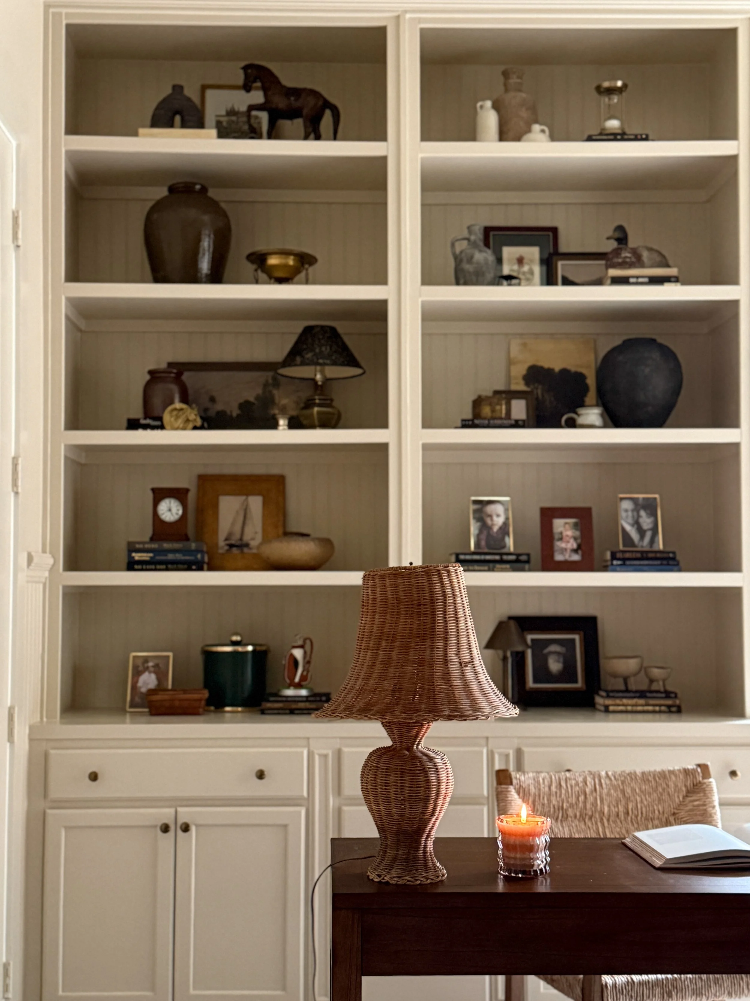 A white bookshelf with decorative objects such as vases, framed photos, books, and sculptures. A part of a wooden table with a rattan table lamp, a lit candle, and an open book is visible in the foreground.