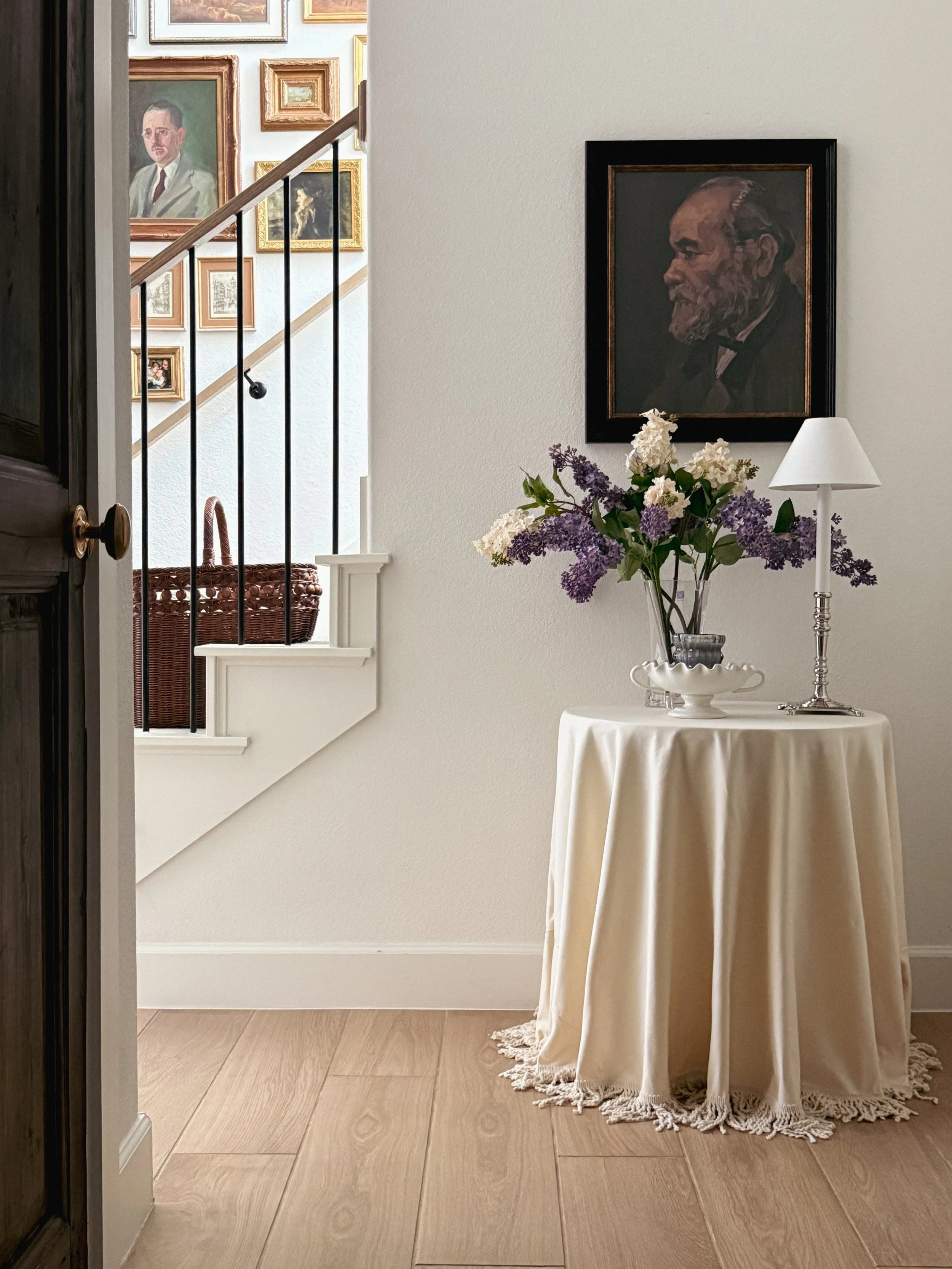 A cozy interior corner with a round table draped in a beige cloth, topped with a glass vase of white and purple flowers and a white lamp, against a plain white wall with a framed profile portrait of an older man with a beard. Behind, there is a stair