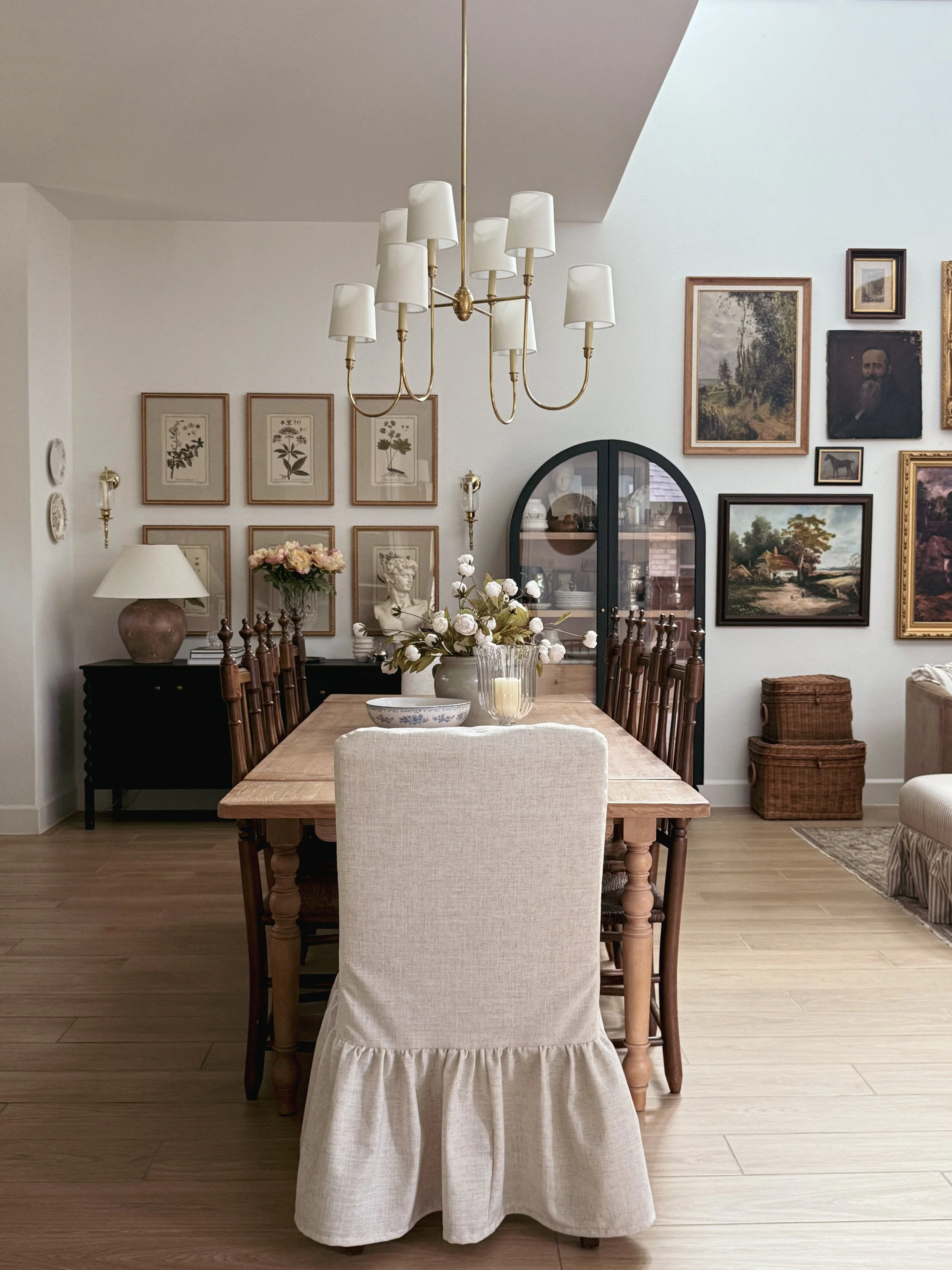 Dining room with a wooden table, white upholstered chair with a ruffled skirt, a black sideboard, and framed artwork on the wall. Decor includes a vase of white flowers, a bowl, and a table lamp. A chandelier with white shades hangs above the table.