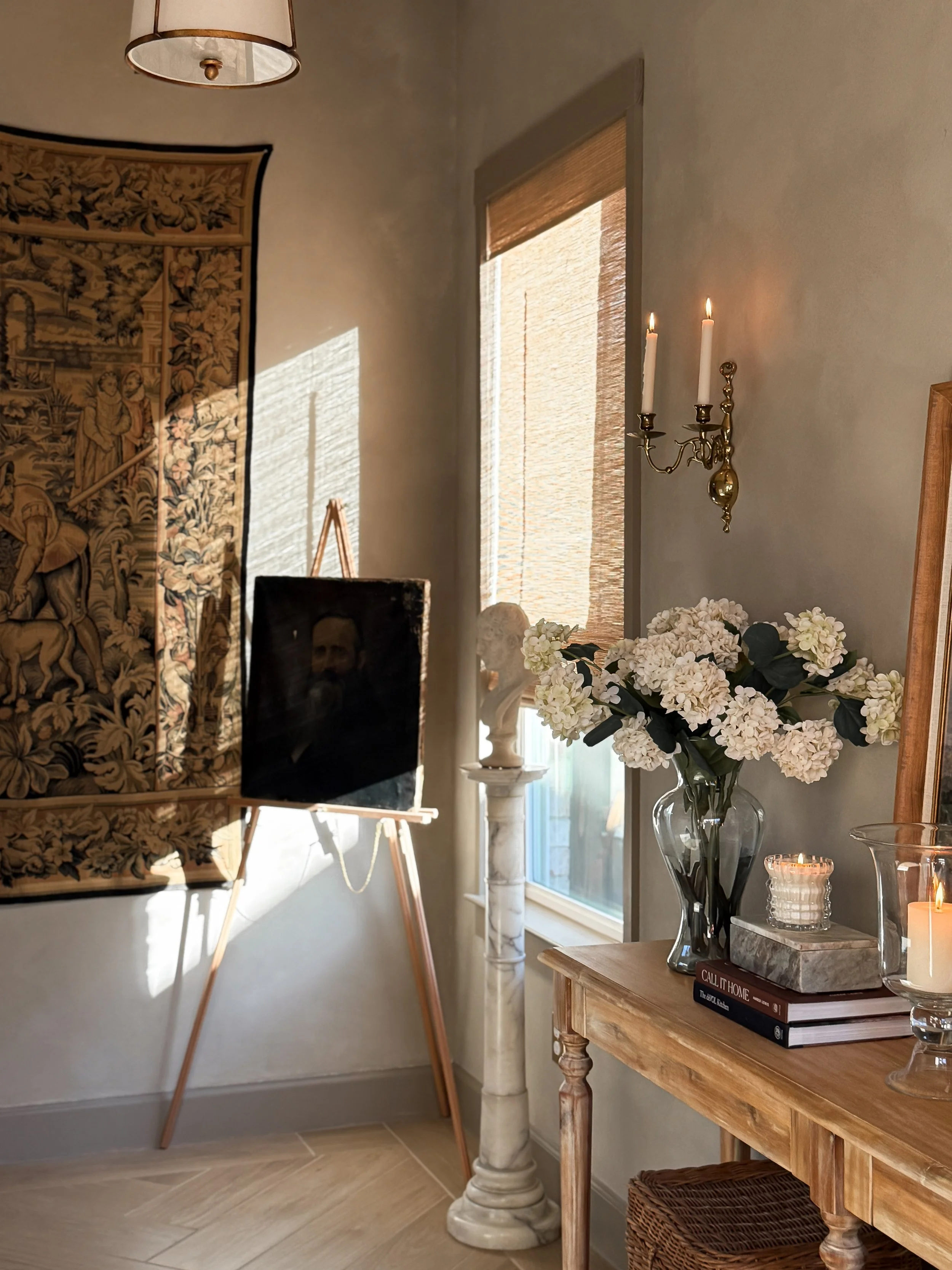 Corner of a room with a table holding white flowers, books, and candles, a wall with a window covered by blinds, a hanging lampshade, and a tapestry on the wall.