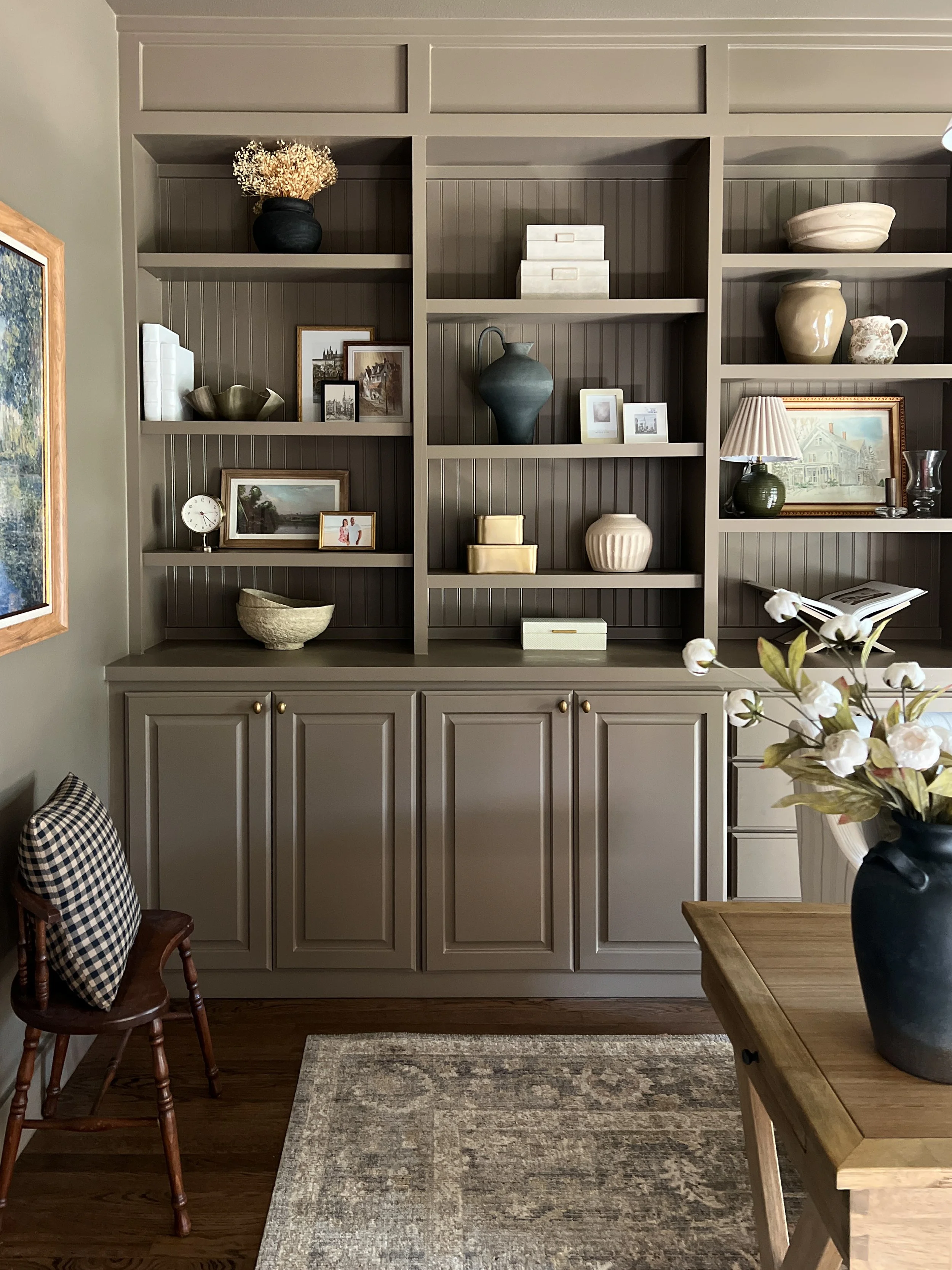 A living room corner with a built-in gray bookshelf, decorated with various framed photos, vases, and decorative objects, partially visible with a wooden table and a bouquet of white flowers in a black vase in the foreground.