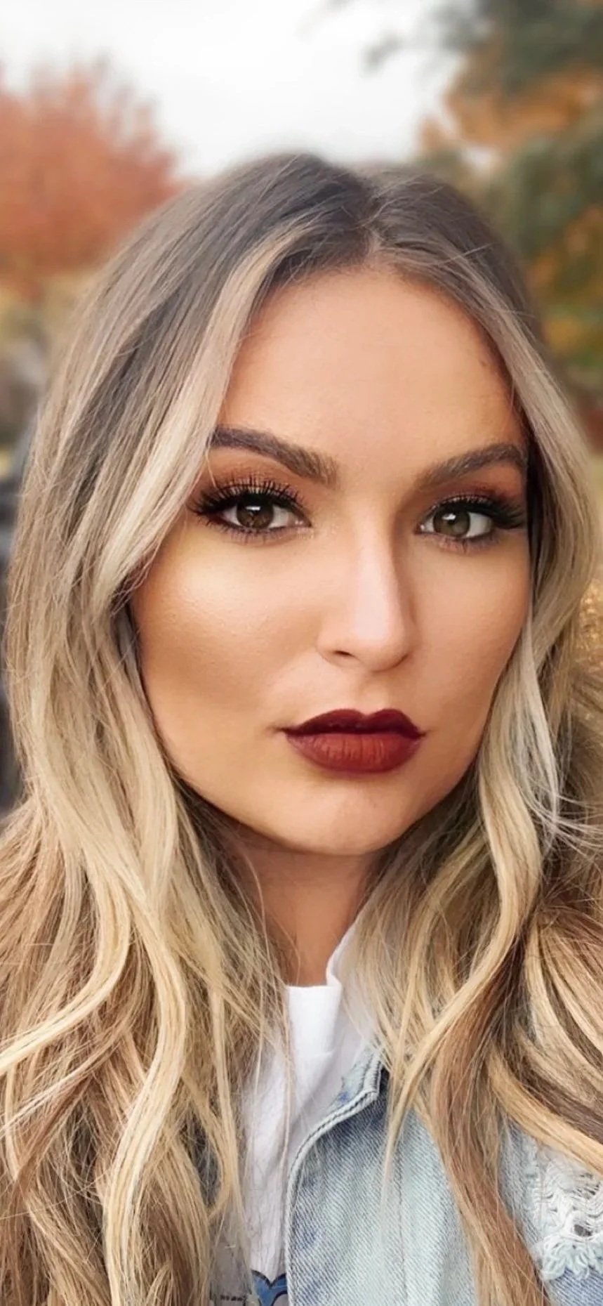 A young woman with long, wavy blonde hair and makeup with dark red lipstick and eye makeup. She is outdoors with autumn trees in the background.