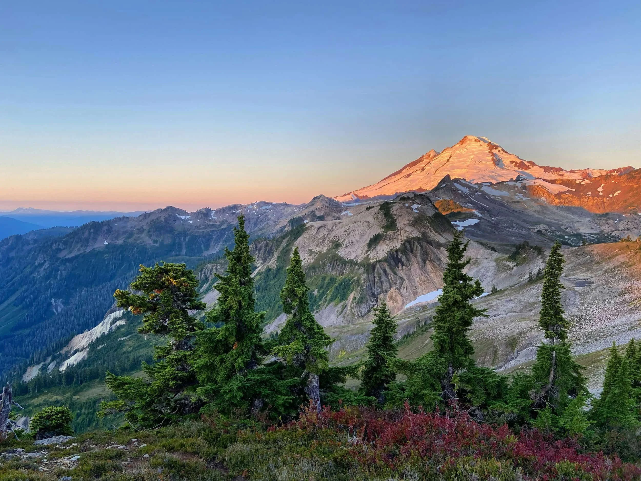 Snow-capped mountain at sunrise with green pines, rugged hills, and clear blue sky—perfect serenity for online therapy in Washington State.