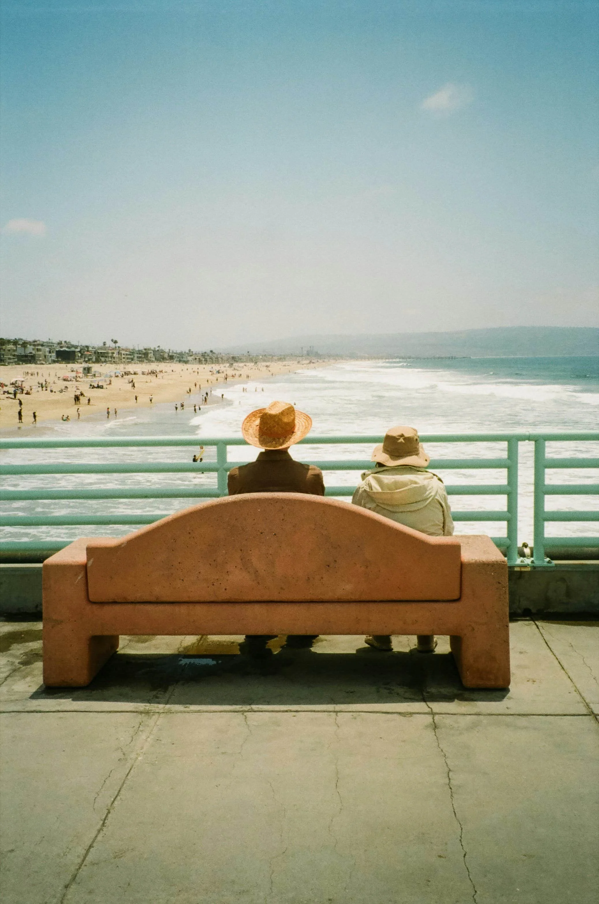 Two people in hats sit on a bench facing the ocean, sharing quiet connection like attachment therapy, overlooking a busy beach.