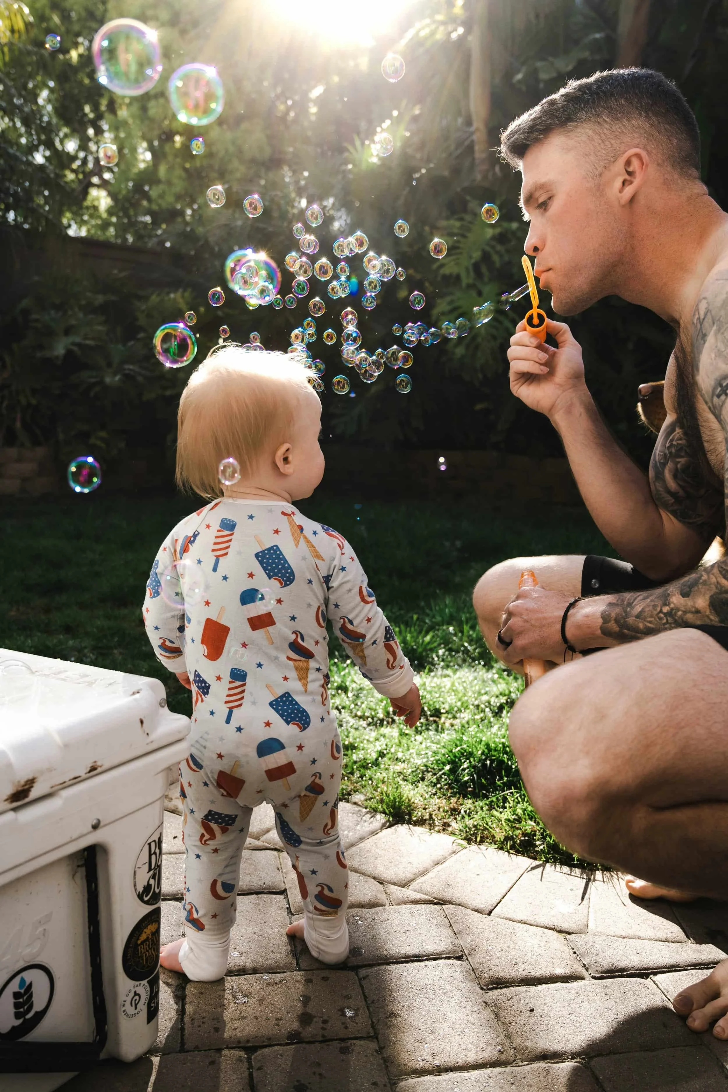 Tattooed man, enjoying therapy for men, blows bubbles towards a toddler in pajamas outside, sunlight shining through the trees.