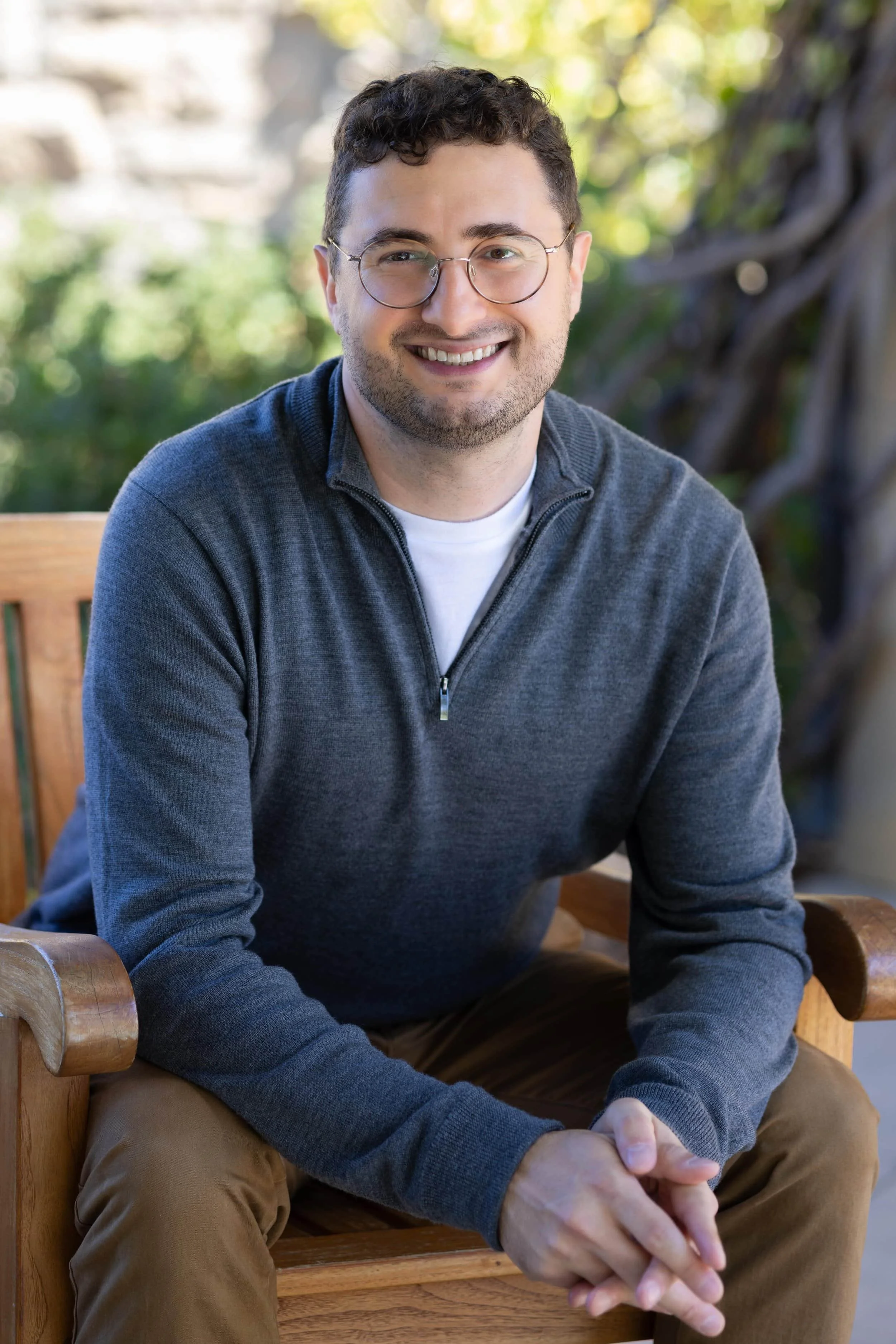 Smiling mental health counselor with glasses, a depression therapist, sits on a wooden bench outdoors in a gray sweater, greenery in the background.