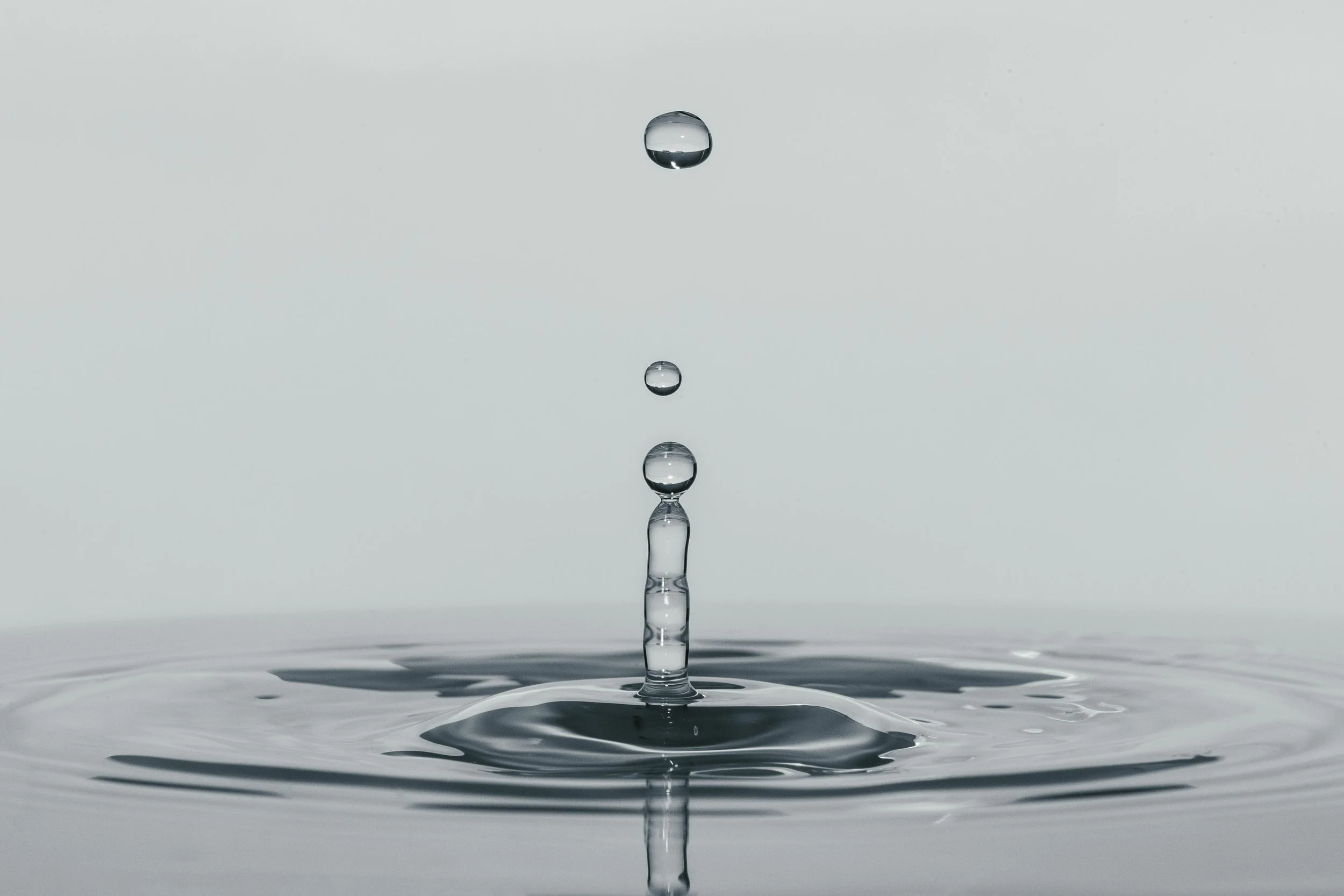 Close-up of water droplets falling into a calm water surface, creating ripples and a vertical column of water.