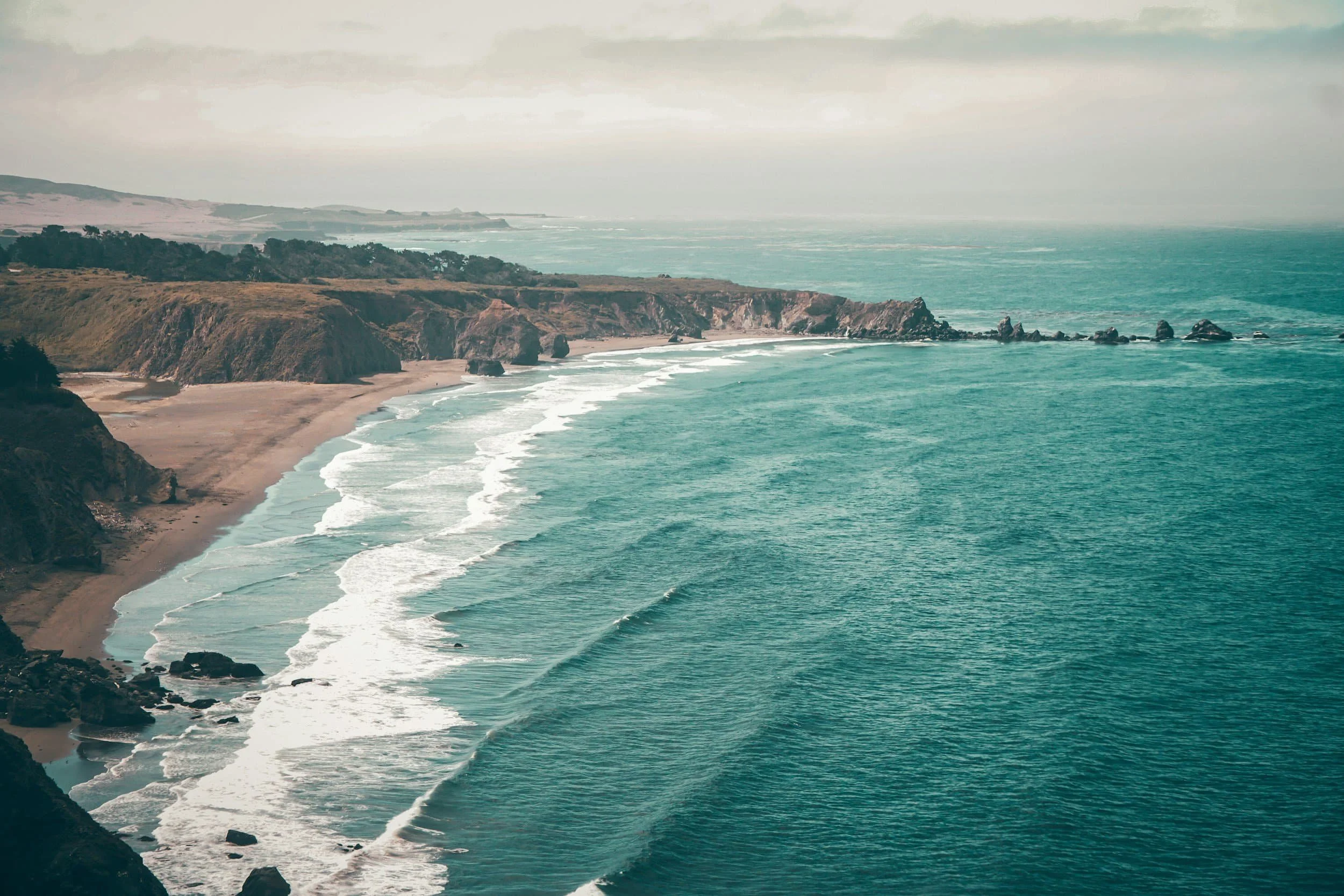 A rugged coastline with gentle waves rolling onto a sandy beach under a cloudy sky, like Online Therapy in the Bay Area—calm and reassuring.