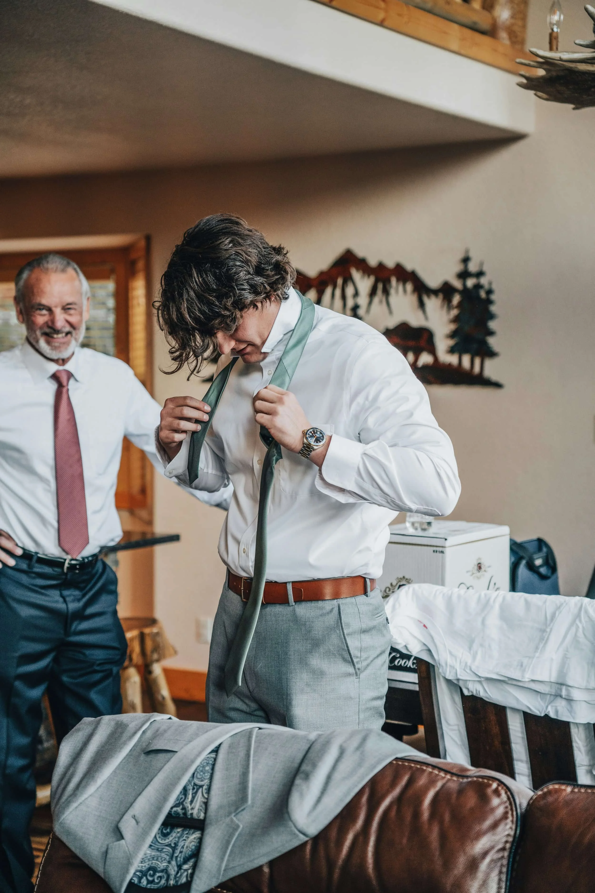 A young man in a white shirt and tie gets dressed as an older man smiles nearby, highlighting therapy for men in a cozy, rustic room.