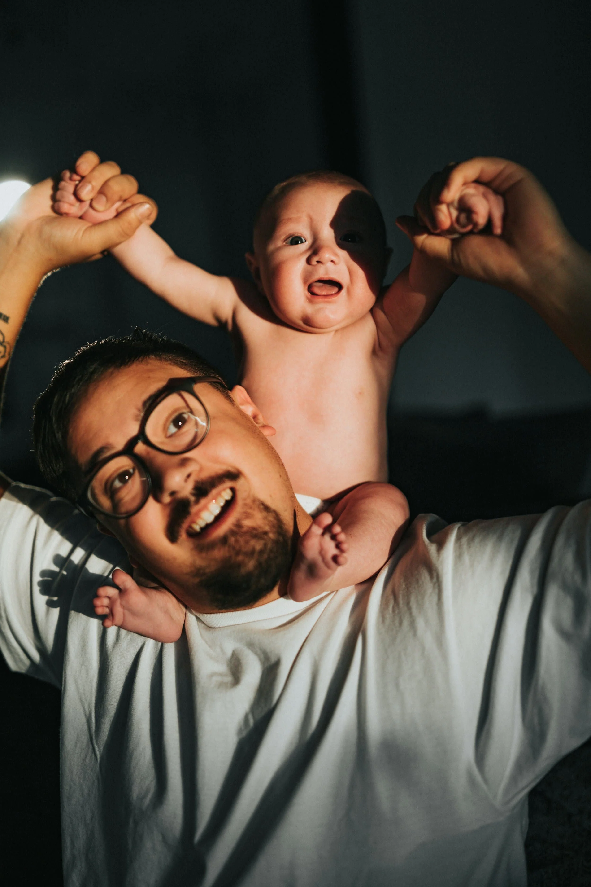 Smiling man with glasses holds a happy baby on his shoulders, both looking up with joy in a warmly lit room—moments of therapy for men.