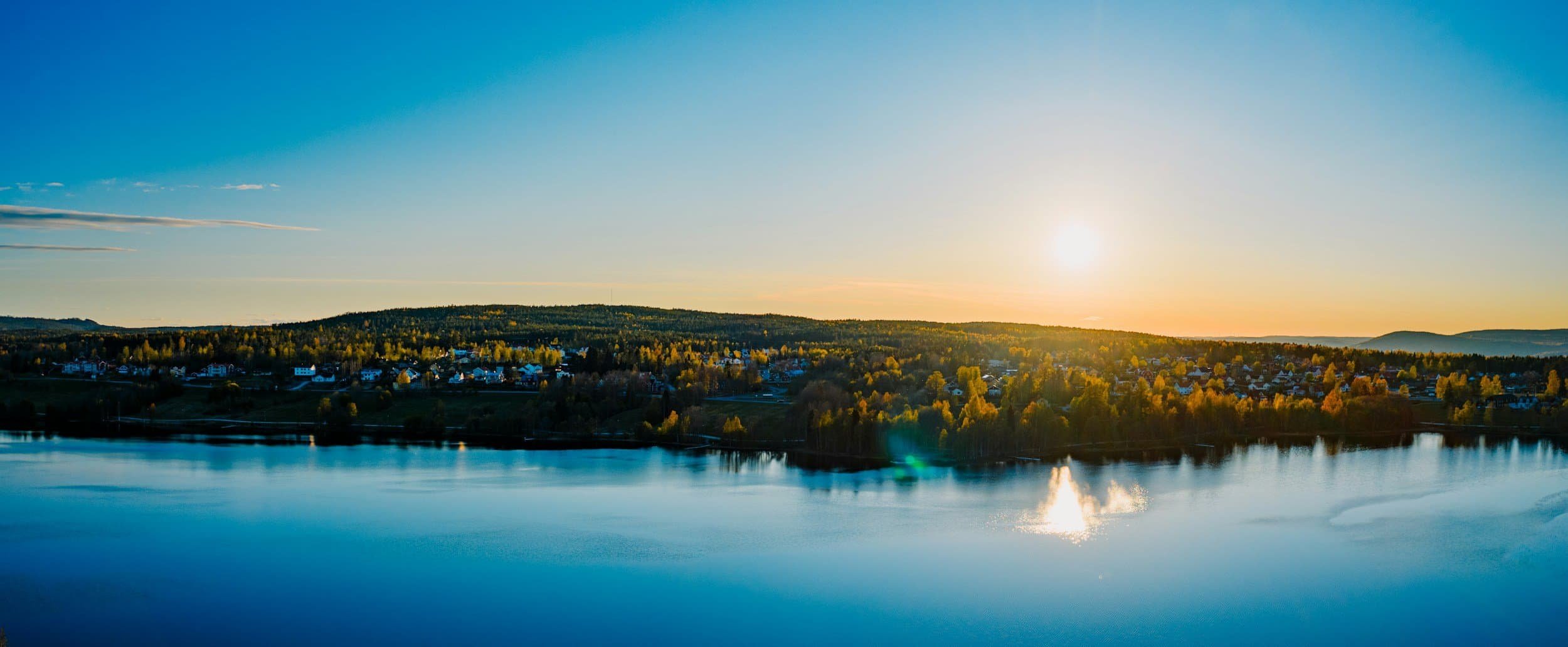 Sunset over a lake with a distant view of a small town and green trees, reflecting on the water.
