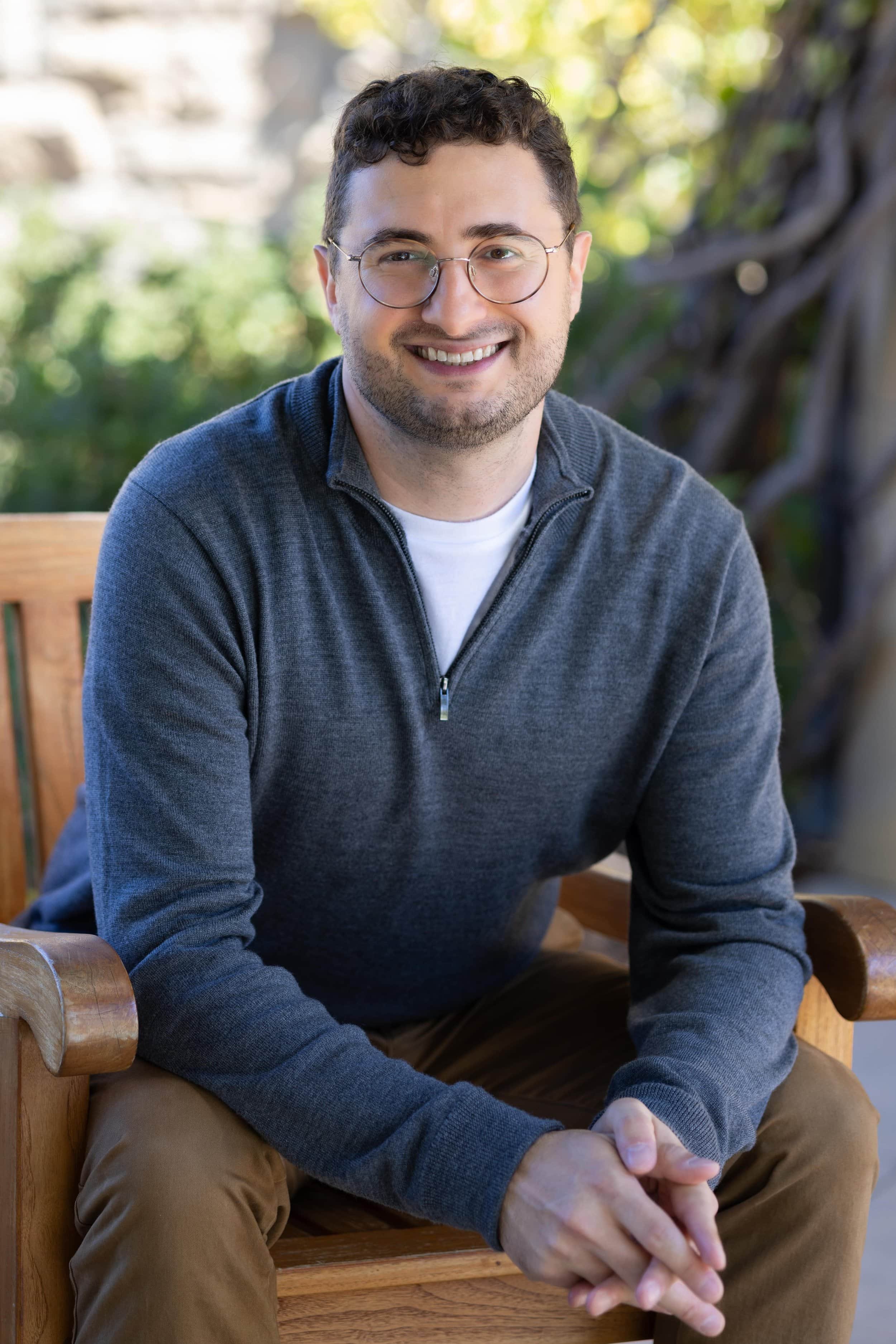Smiling man with glasses sits on a bench outdoors, wearing a gray sweater—perhaps reflecting after relationship therapy, hands clasped.