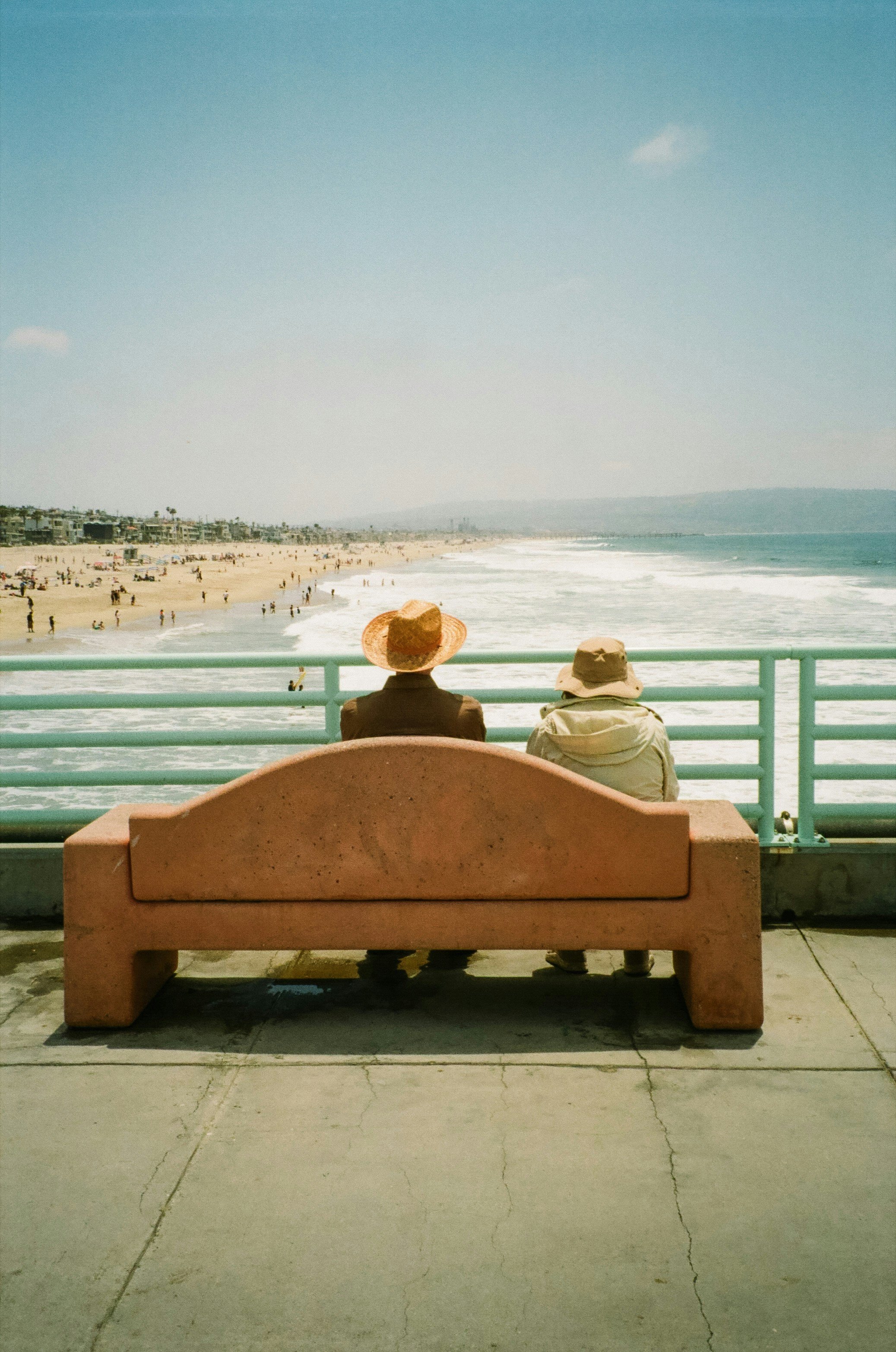 Two people sitting on a pink bench at a pier, facing the ocean, with a beach and many people in the background, under a clear blue sky.