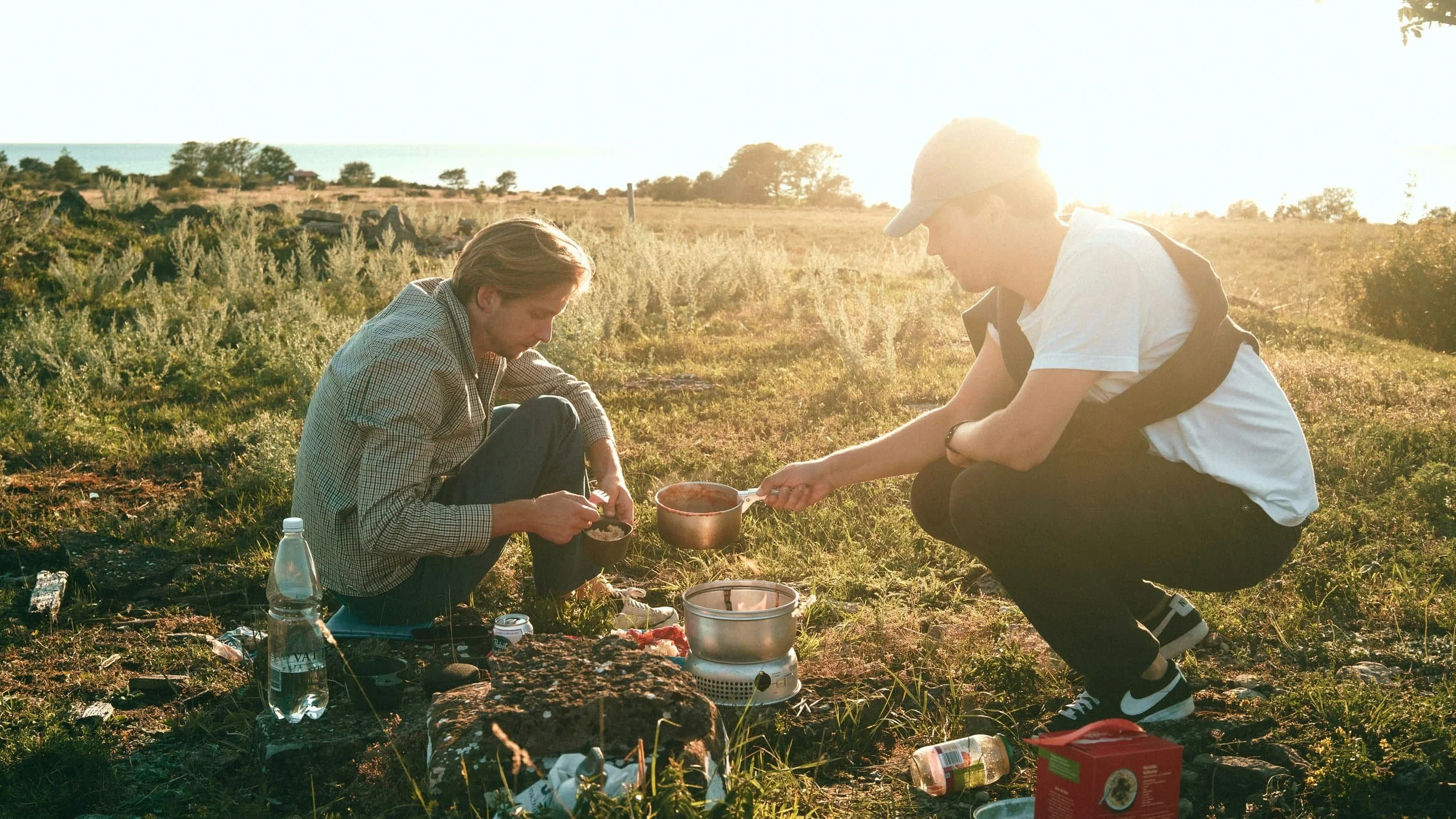 Two people cooking outdoors at sunset, kneeling by a stove—outdoor therapy for men surrounded by camping supplies.