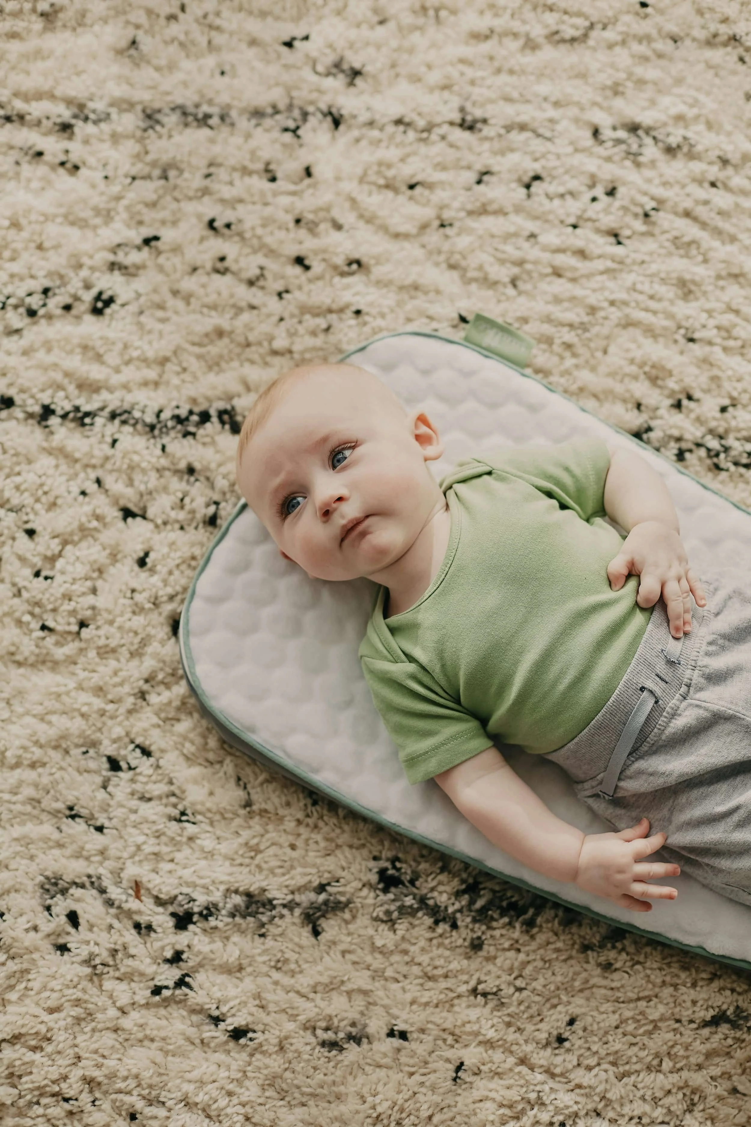 A baby in a green shirt and gray pants lies on a mat, an ideal setting for attachment therapy, gazing off to the side.