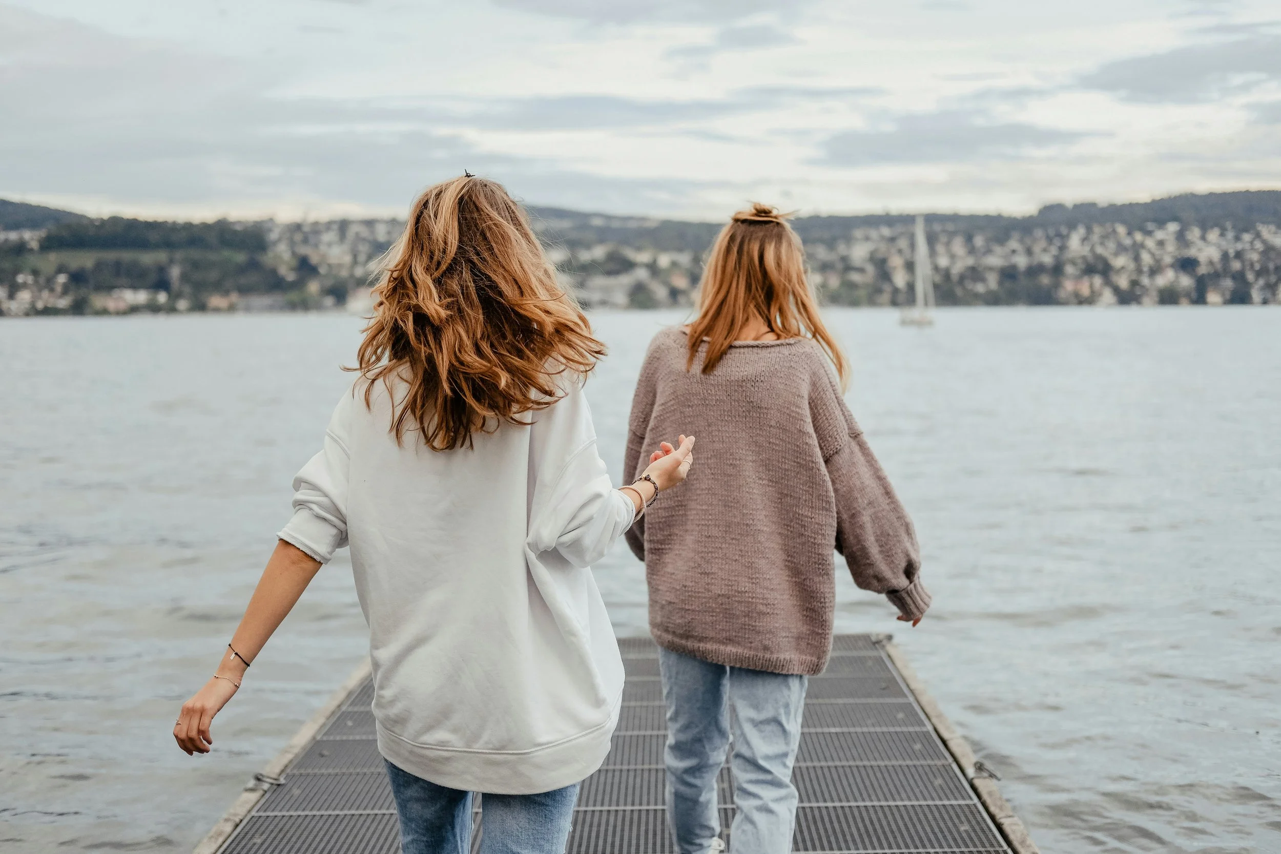 Two people with long hair walk along a dock toward a lake, as clouds gather—a tranquil scene evoking depth therapy’s calm introspection.