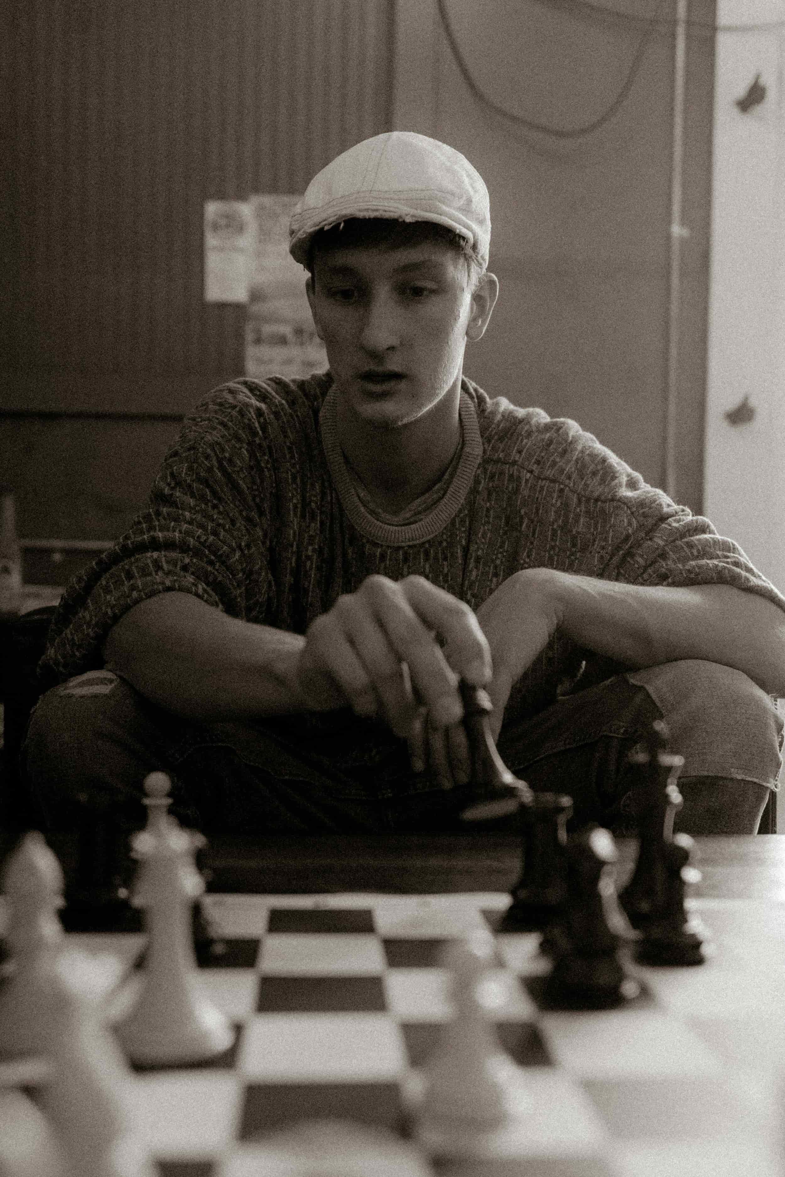 A young man in a cap and sweater sits, focused, as he plays chess, finding calm through therapy for men.
