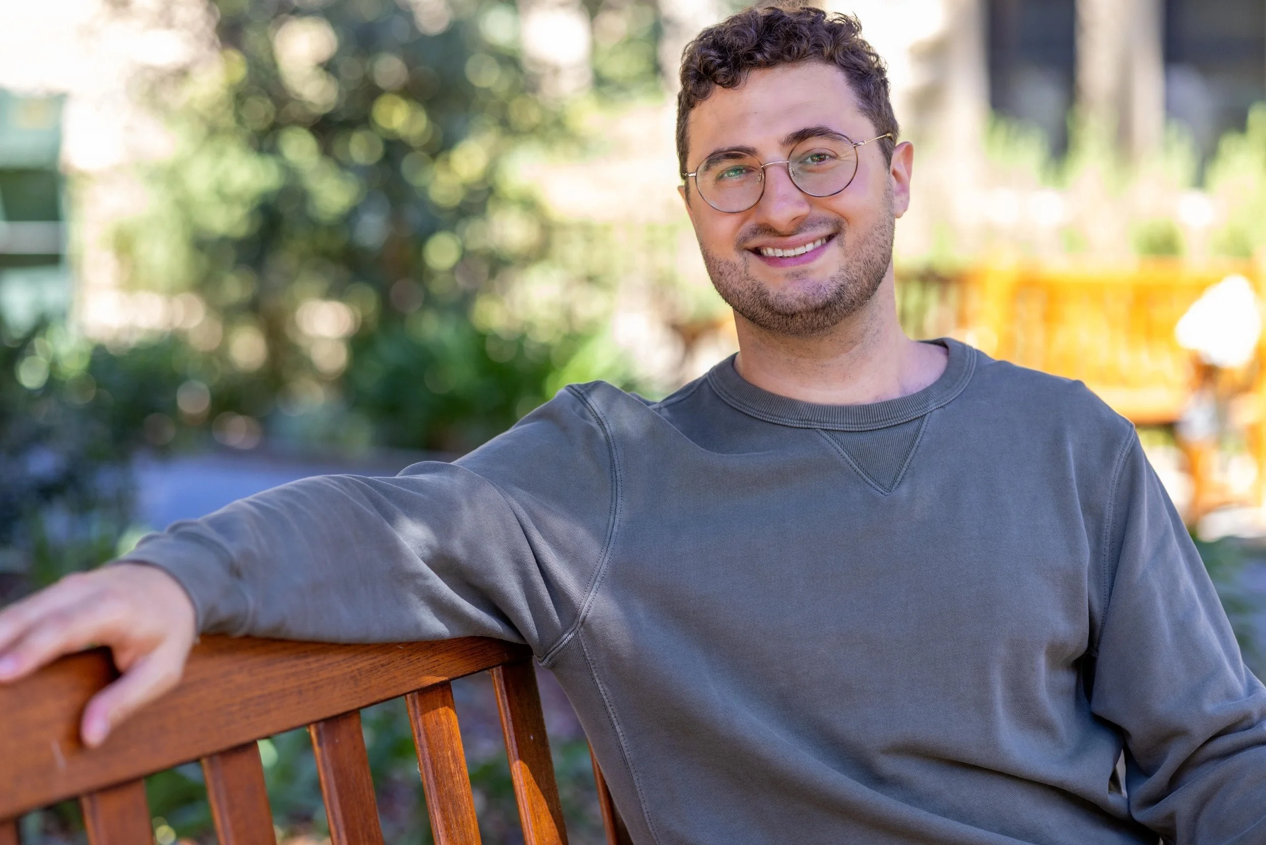 A trauma therapist with curly hair and glasses smiles while sitting on a wooden bench outdoors.