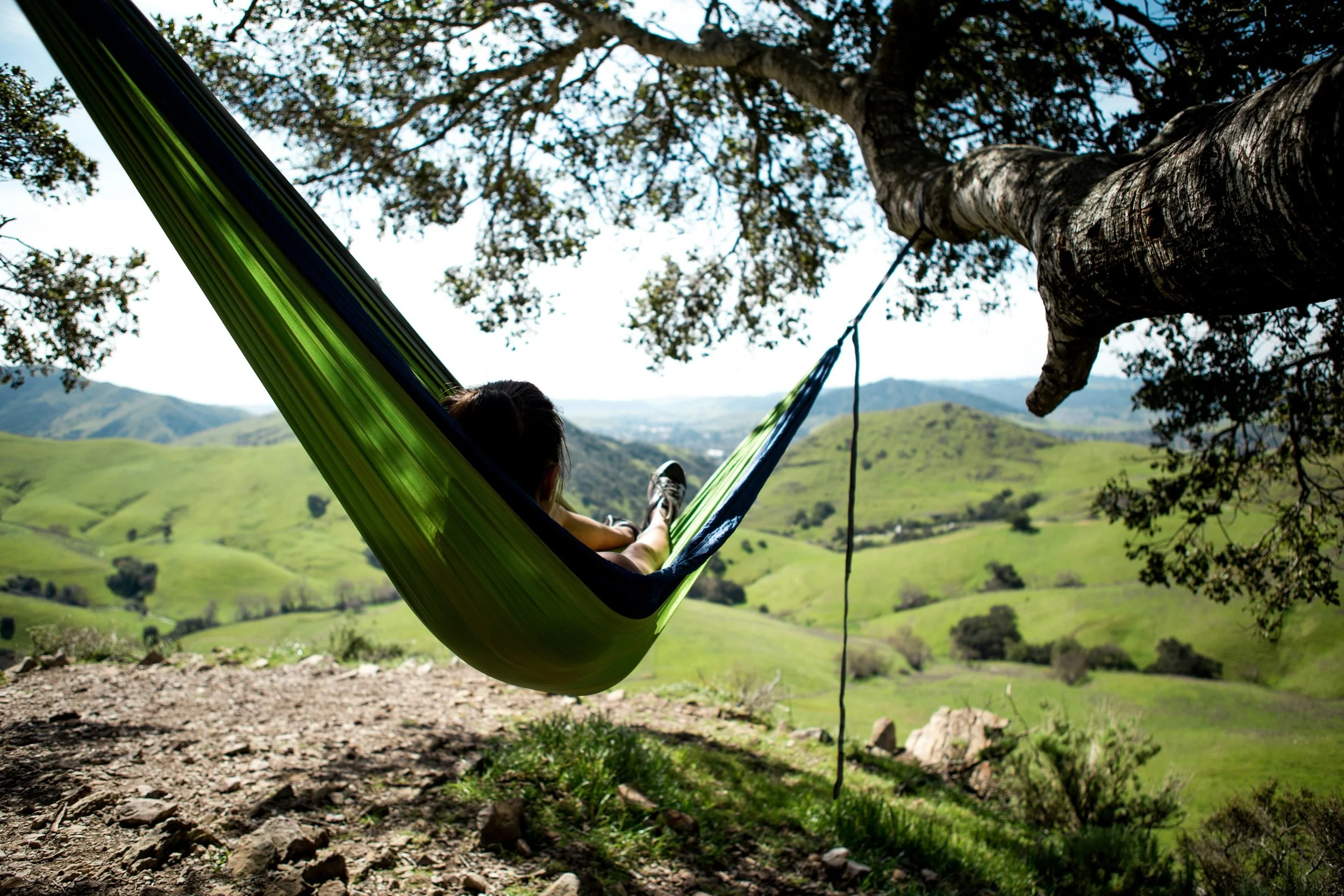 Person lying on a green hammock tied to a tree, overlooking rolling green hills in a scenic countryside on a sunny day.