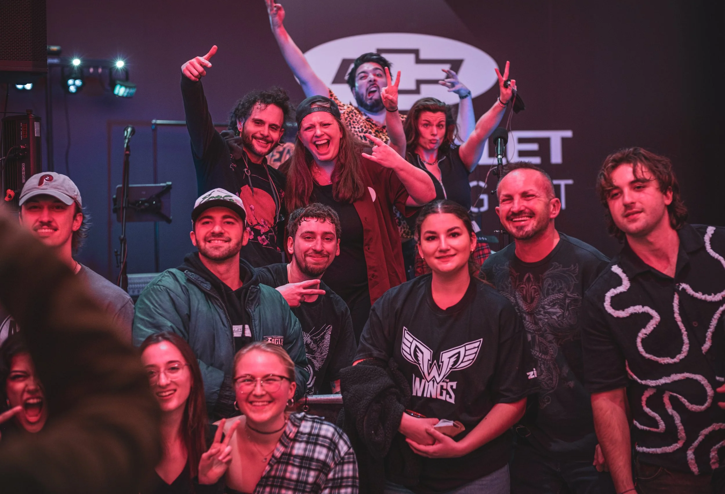 Group of people posing on stage at a concert or event, some making peace signs, with a large logo and text in the background.