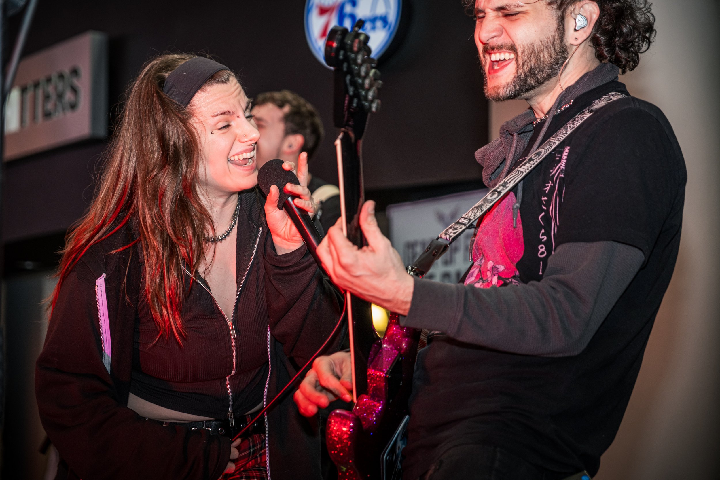 A woman singing into a microphone while a man plays guitar, both sharing a joyful moment during a live music performance.