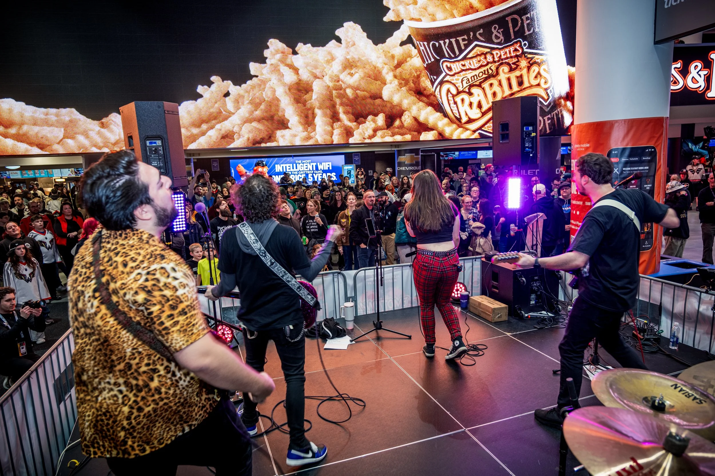 A band performing on a stage at a busy event with a large crowd watching. The background features a giant screen displaying an image of popcorn, a cup of Raisin Bran cereal, and a Chevrolet sign.
