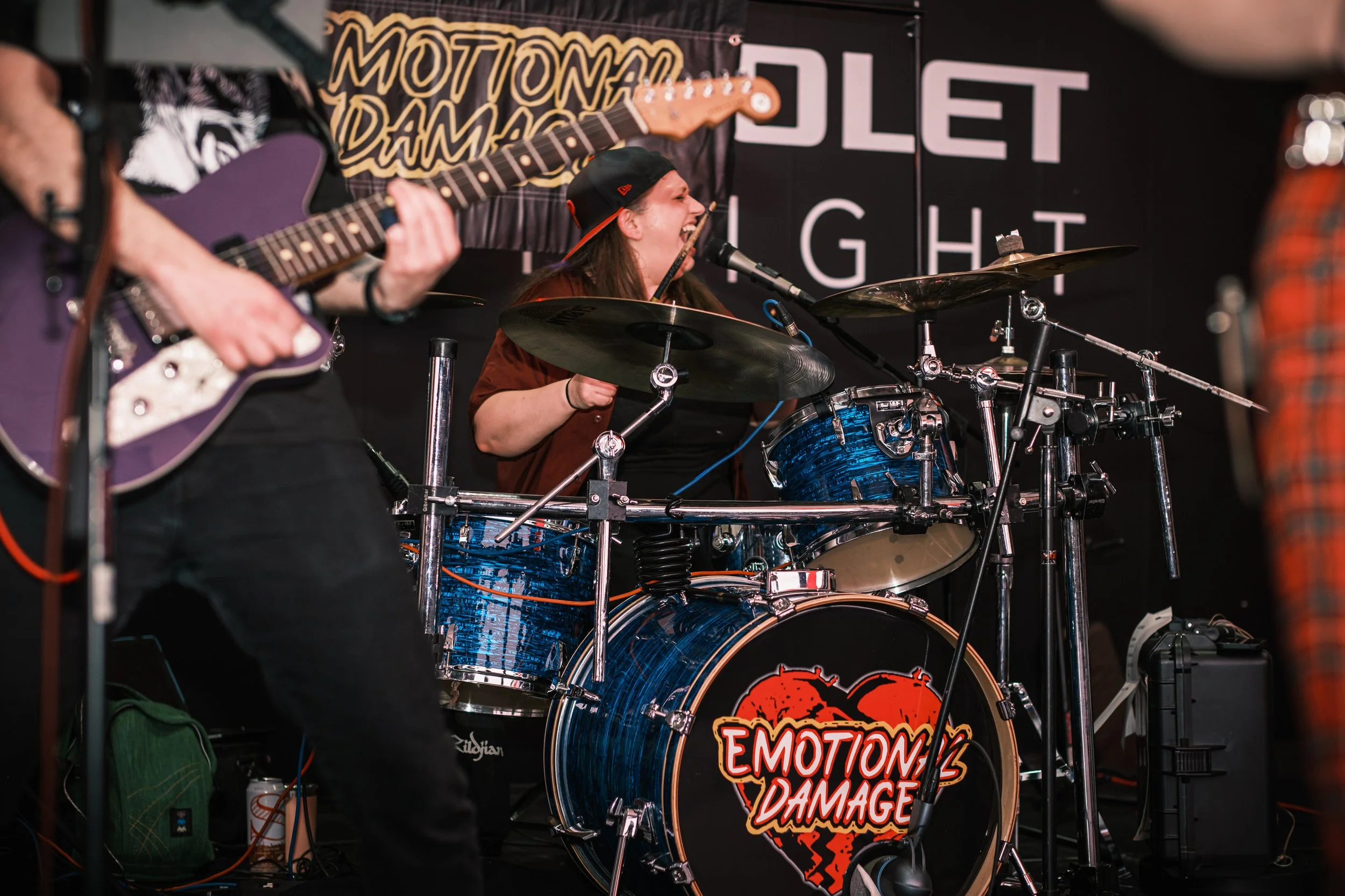 A woman playing drums with a racing stripe-style drum kit featuring a heart graphic and the words 'Emotion Damage' on the bass drum, singing into a microphone while a guitarist plays nearby.