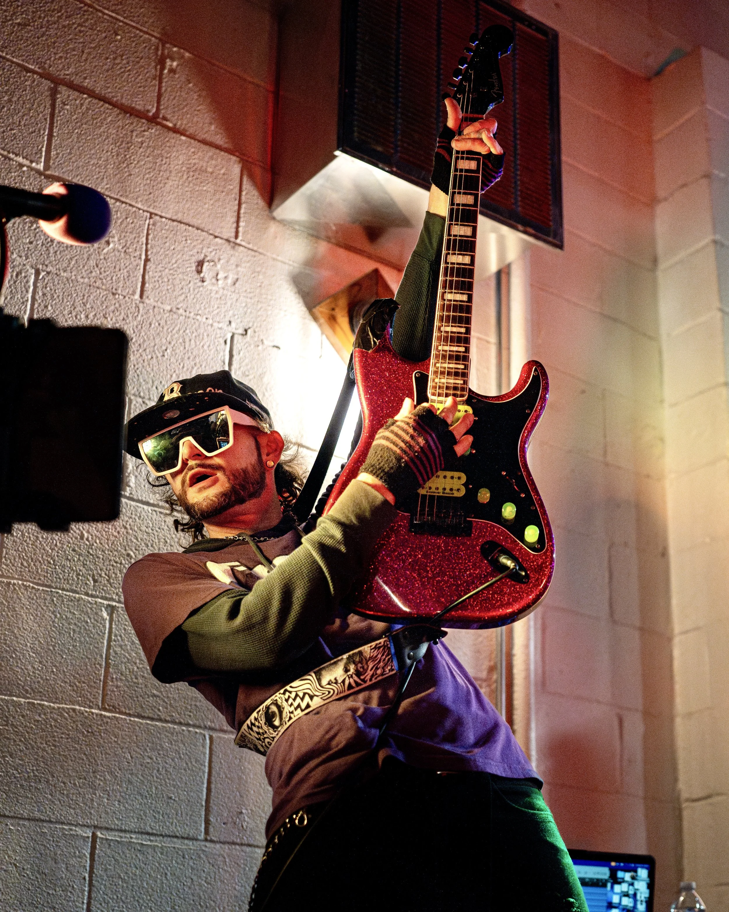 A man wearing sunglasses and a cap plays an electric guitar on stage, with colorful lighting and a textured brick wall in the background.