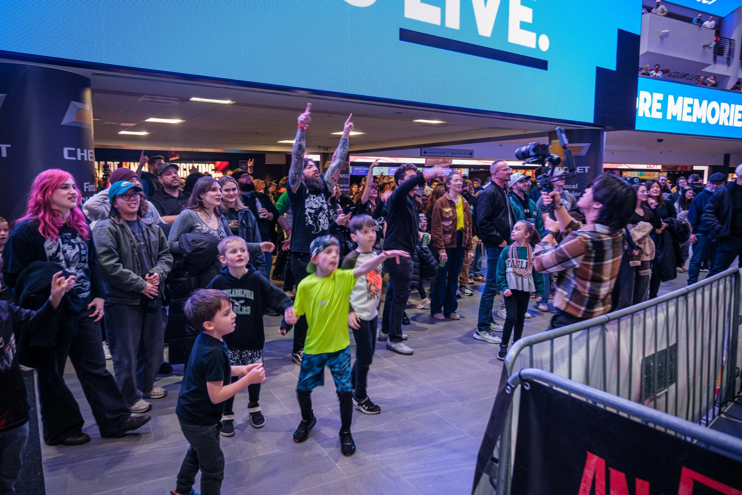 A group of people, including children and adults, at a public event in an indoor venue, watching a performer with a camera. The crowd is engaged, some raising their hands, under a large digital screen displaying blue text.