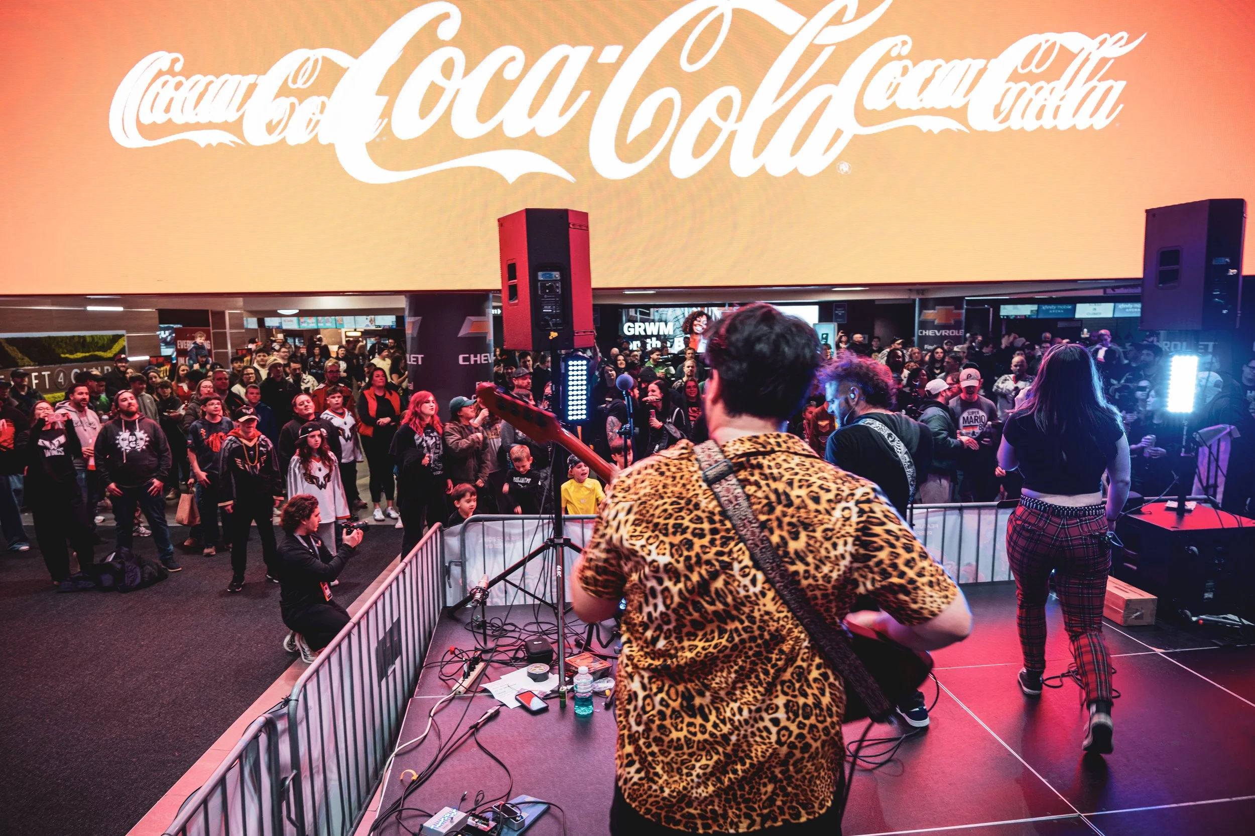 Musicians perform on a stage at an indoor event with a large crowd watching. The stage has a few electric guitars, and there's a large screen with the Coca-Cola logo projected behind the performers. The audience appears engaged, with some taking photos, and some standing near the stage.