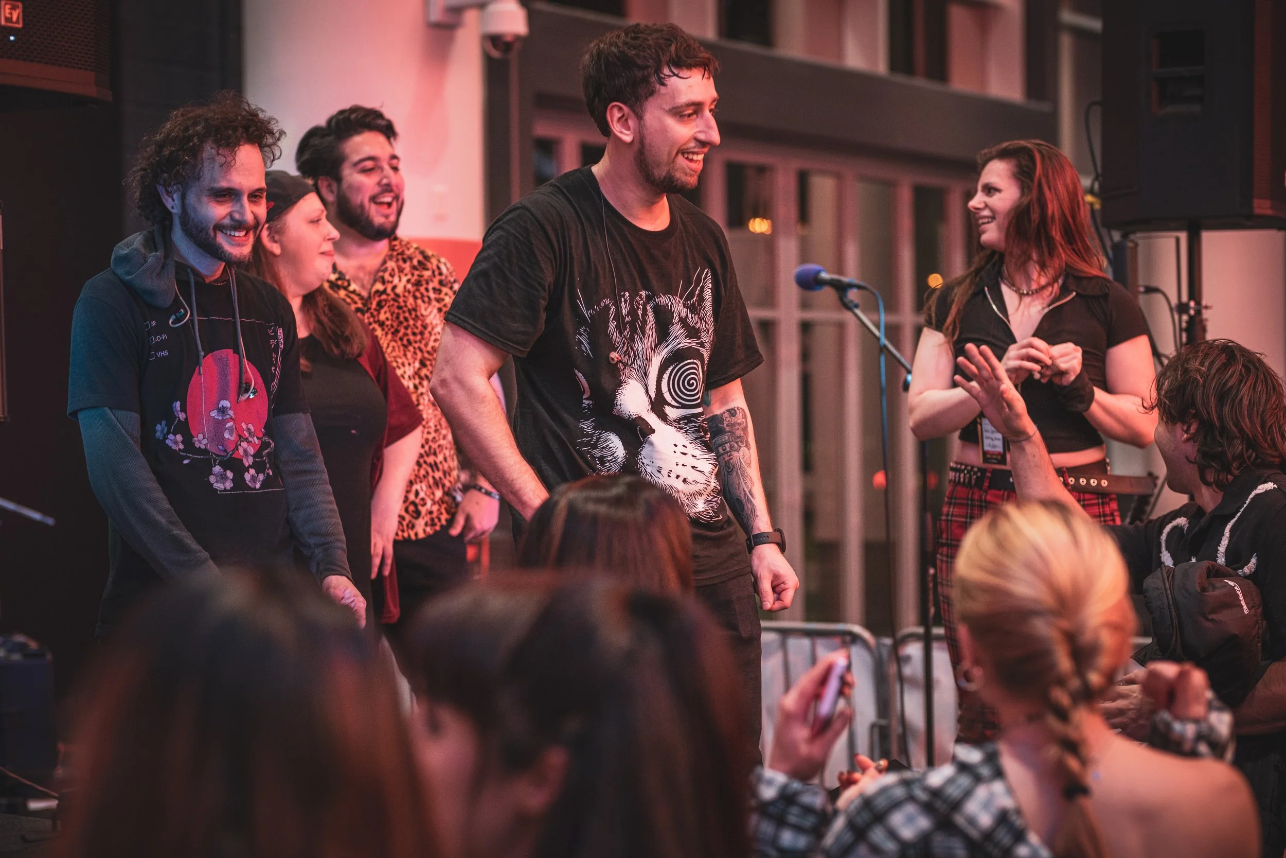 Group of people on stage at a social event, with one person smiling and others clapping or engaging with the crowd, in a lively indoor setting.