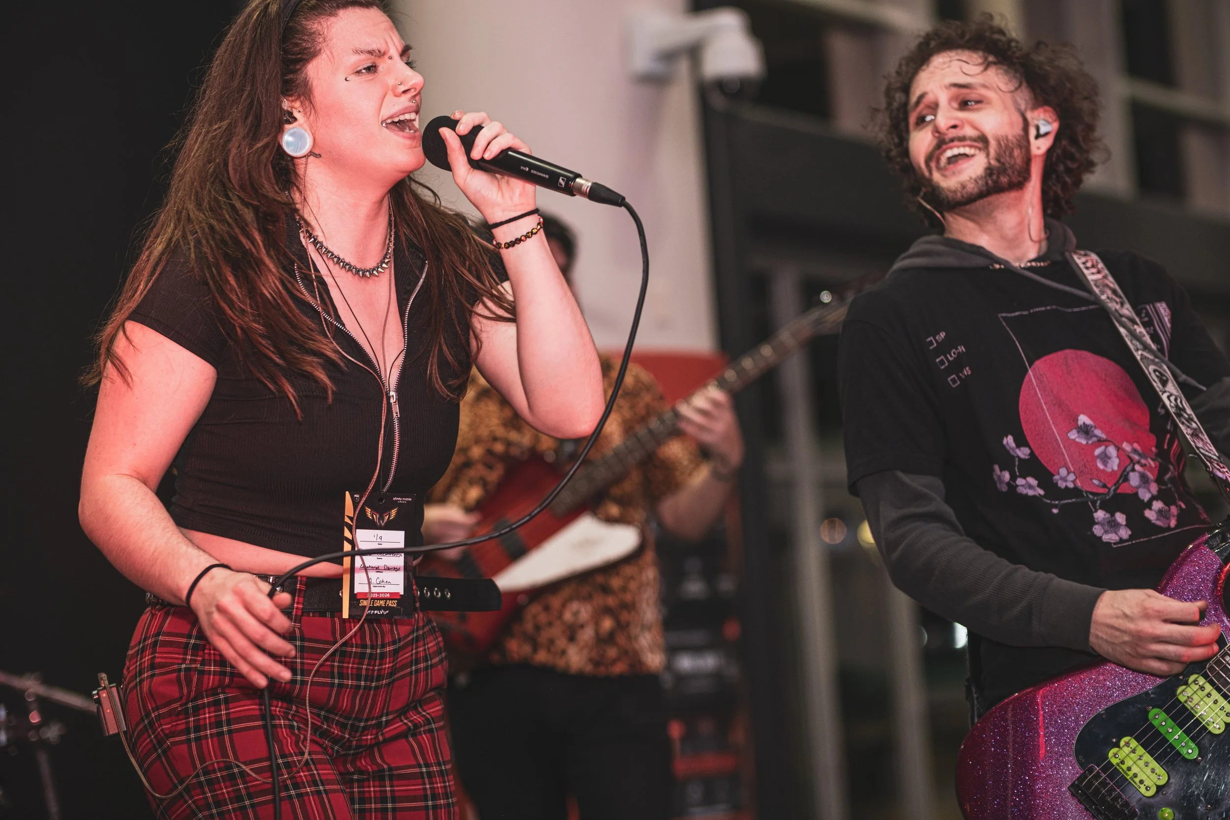 A woman singing into a microphone, wearing red plaid pants and a black cropped zip-up top, with a man beside her playing a glittery purple electric guitar. In the background, another person is playing an acoustic guitar.