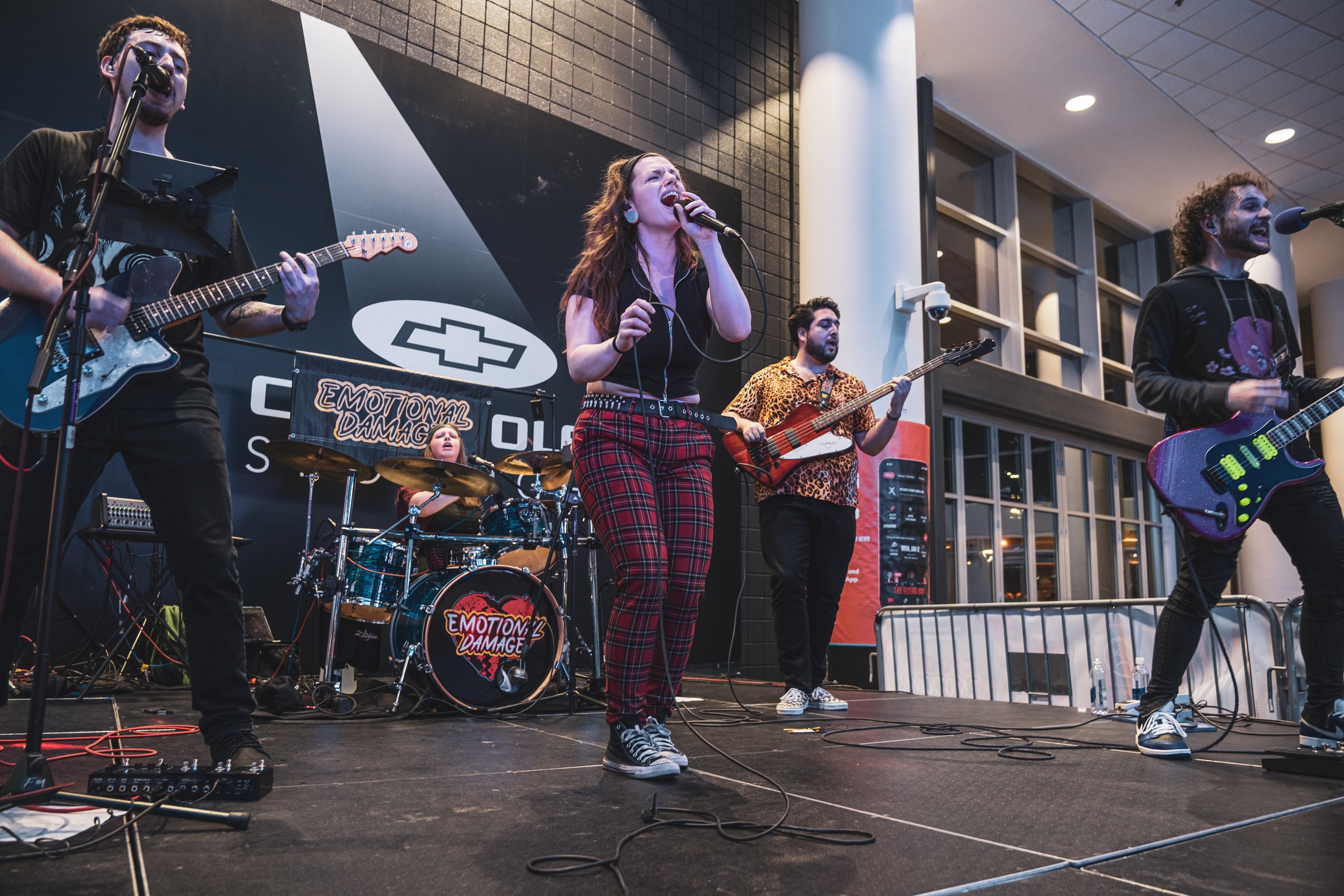 A band of five musicians performing on stage, including a female vocalist, two guitarists, a bassist, and a drummer, with a black backdrop featuring Chevrolet graphics and the words "Emotional Damage."