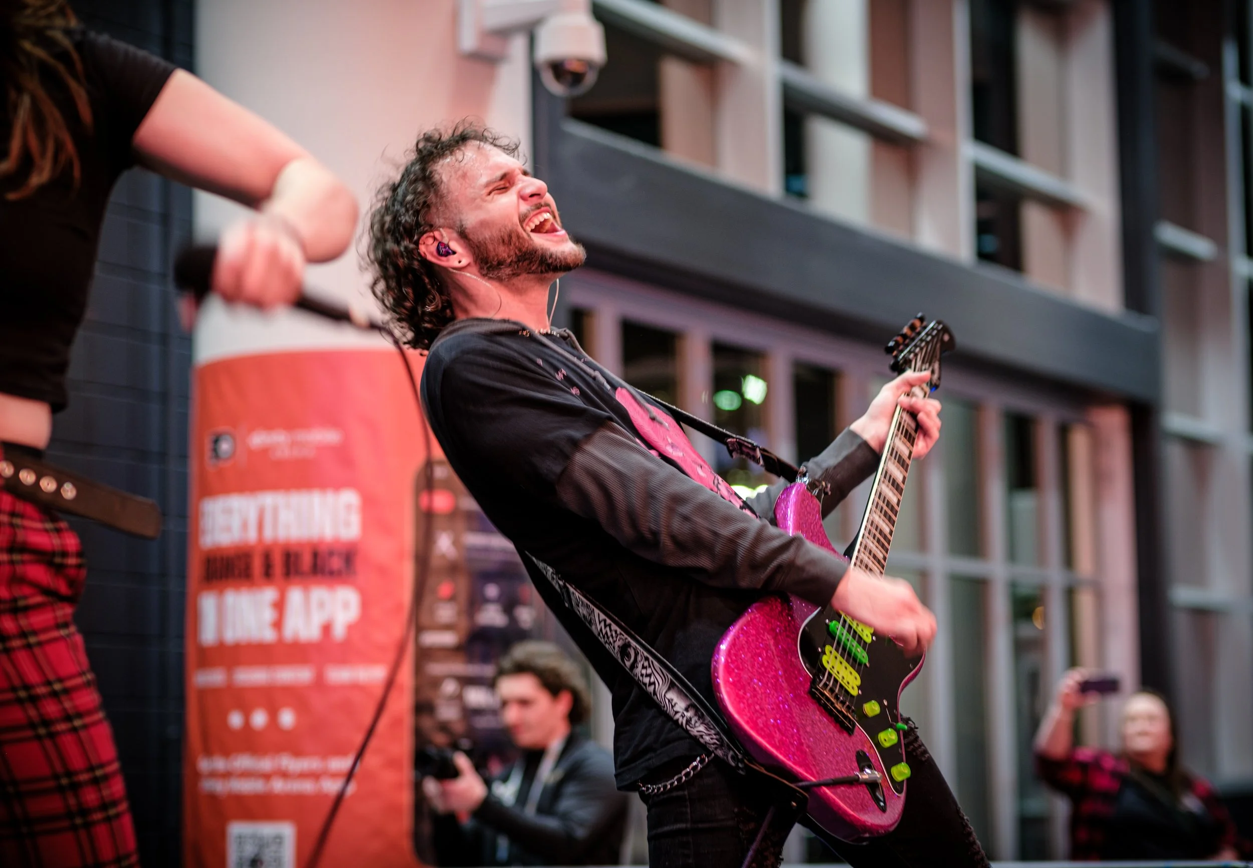 A man with curly hair and a beard playing a sparkly pink electric guitar, performing passionately on stage with an emotional expression.