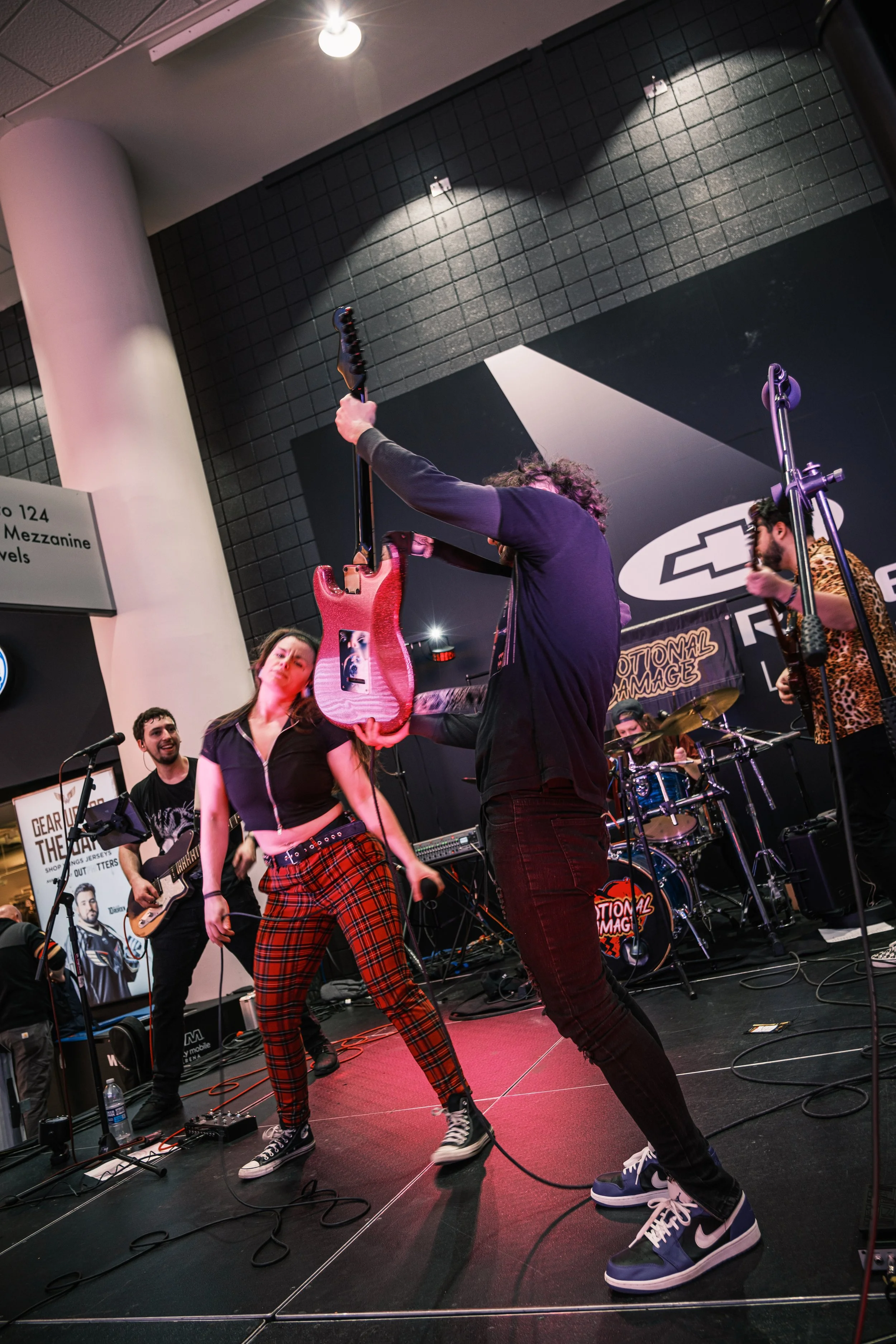Musicians performing on stage, one person holding a guitar above their head, others playing instruments, with a band logo in the background in a concert setting.