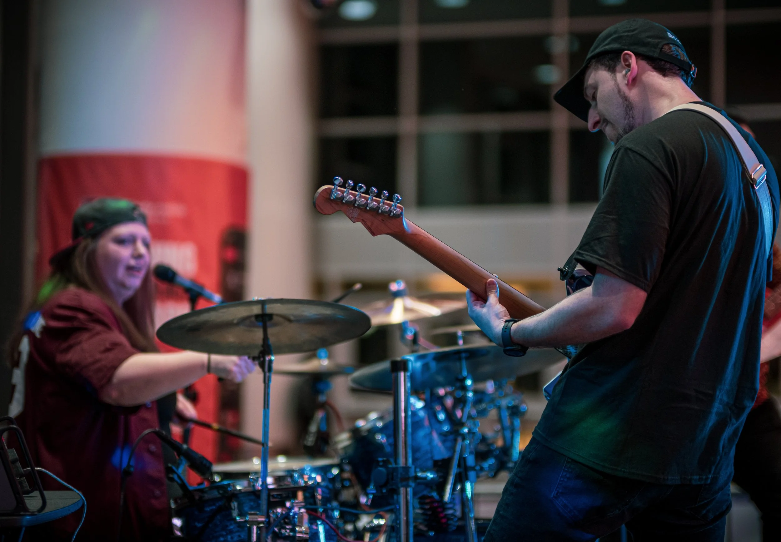 A female musician singing into a microphone while playing drums, and a male musician playing an electric guitar in a band performance setting.