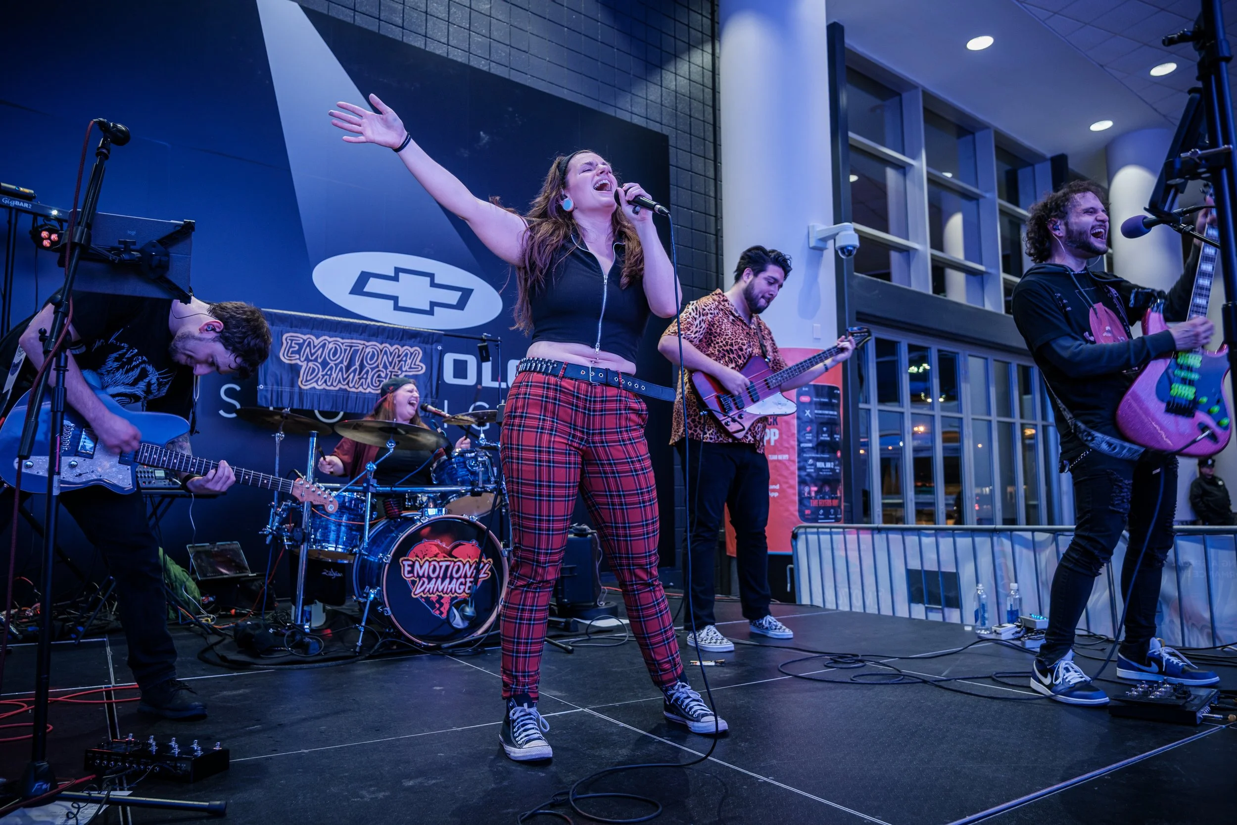 A band called 'Emotional Damage' performs on stage with a female lead singer, three male guitarists, and a drummer in the background inside a modern building with large windows.