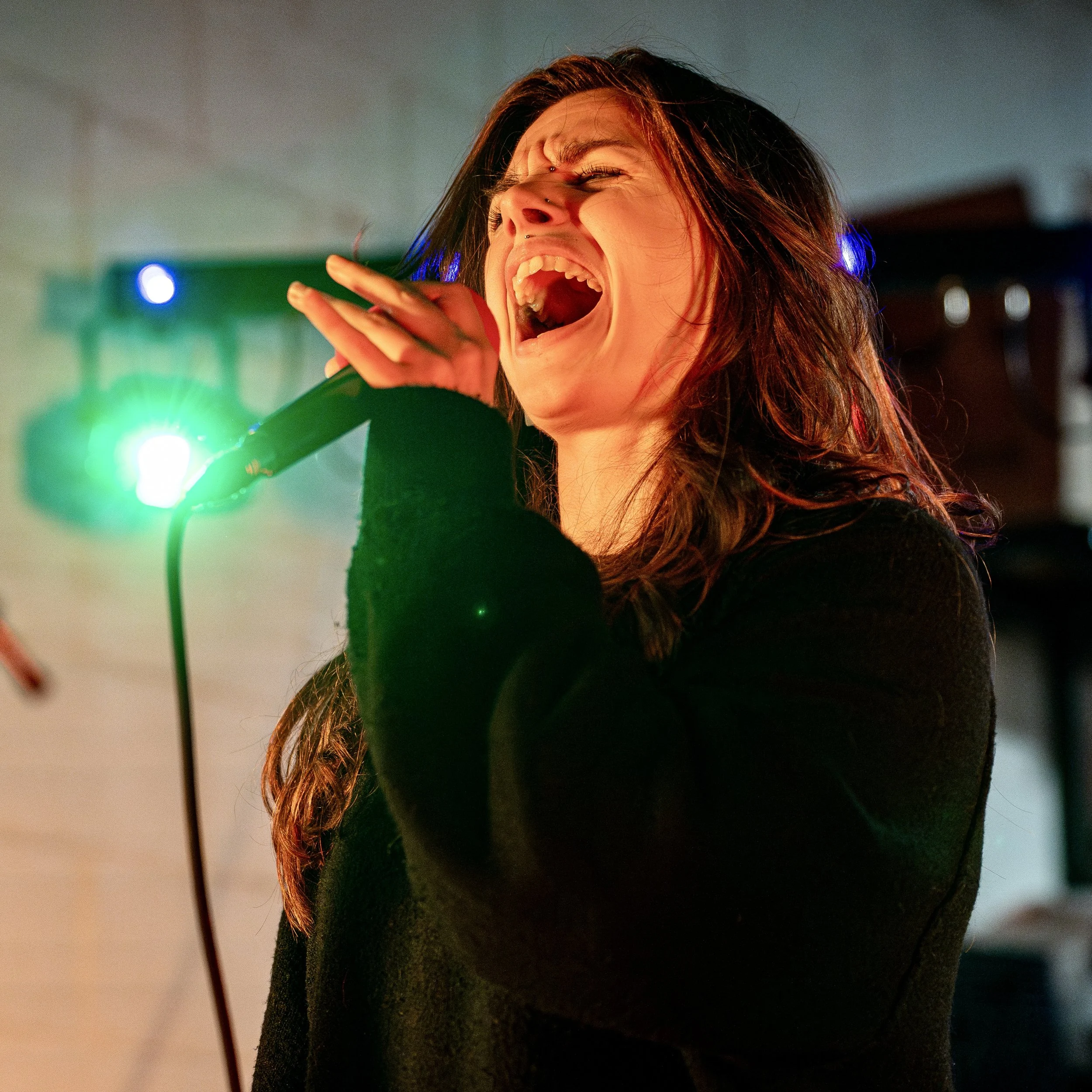 A woman with long brown hair singing into a microphone with green and purple stage lighting in the background.