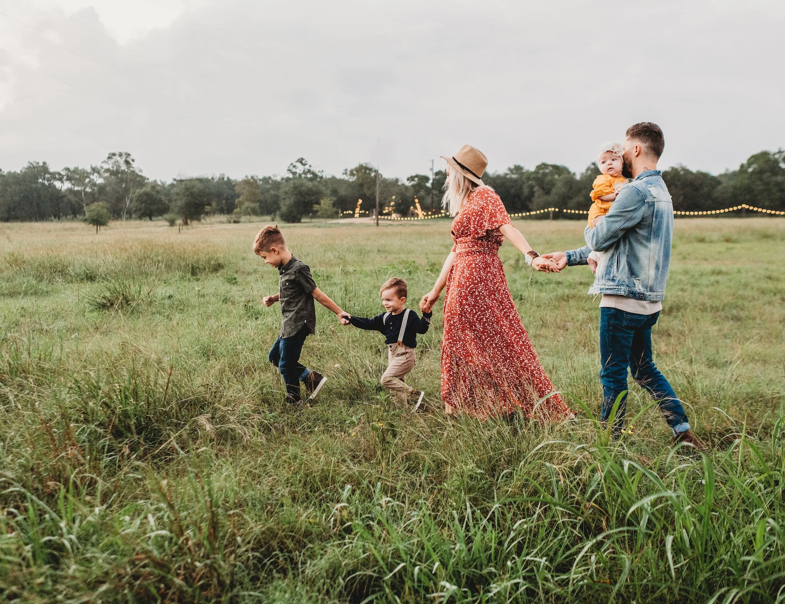 A family of five walks hand in hand through a grassy field, with a cloudy sky in the background. The mother, wearing a floral dress and hat, holds a child's hand, while the father, in a denim jacket, carries a baby. String lights are visible in the distance.