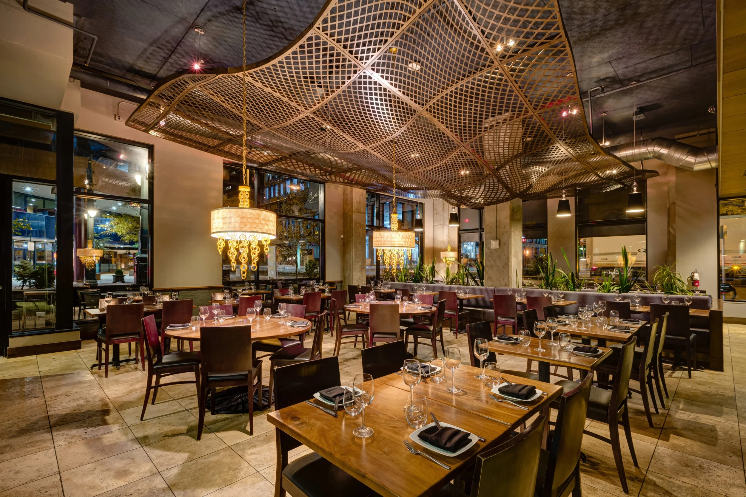 Interior of a modern restaurant with elegant decor, featuring wooden tables set with glassware and napkins, plush seating, and ornate chandeliers. A distinctive lattice ceiling adds to the stylish ambiance.