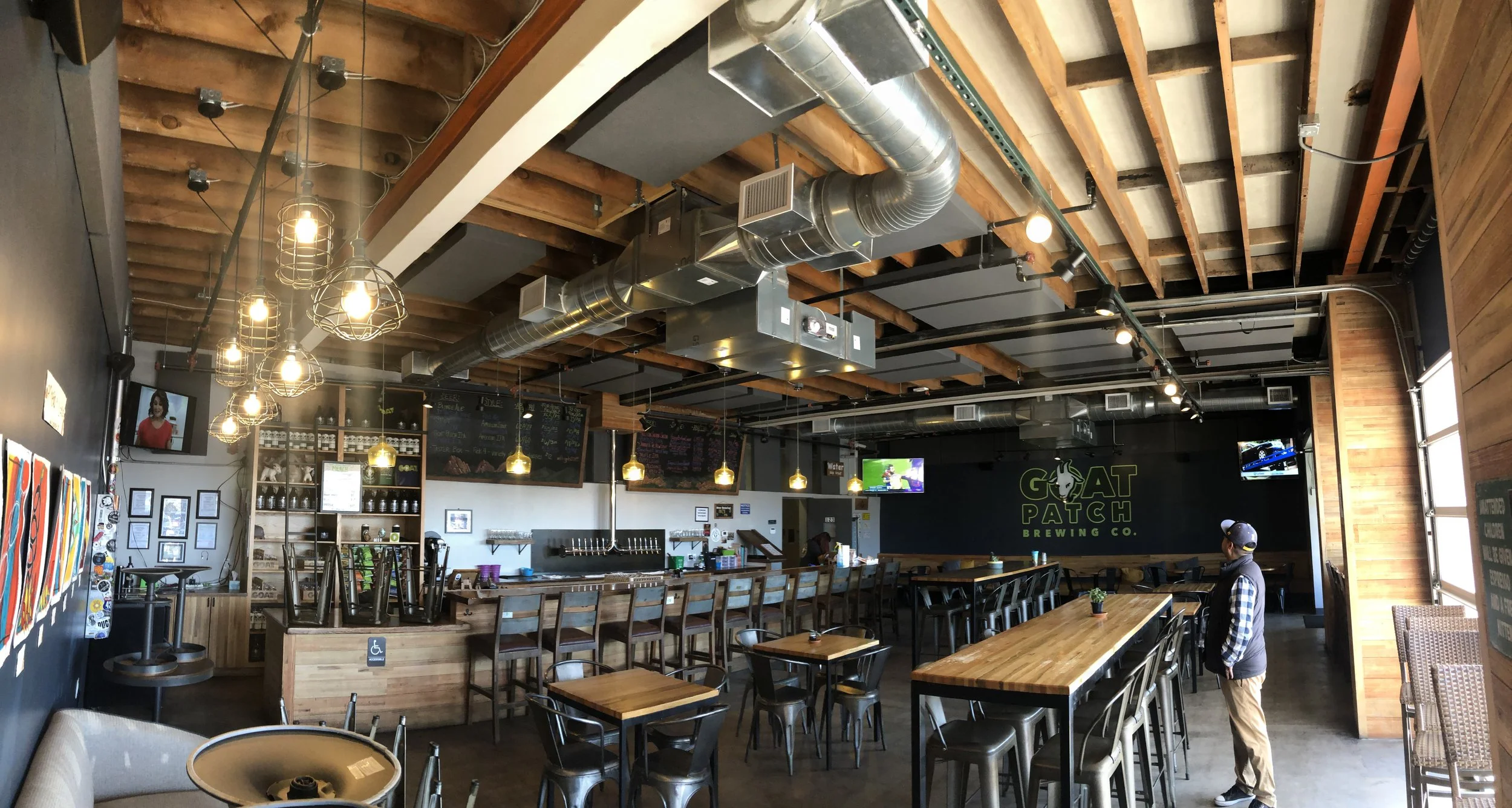 Interior of a brewery taproom with wooden bar, tables, industrial-style lighting, metal ducts, and a person standing near a large communal table.