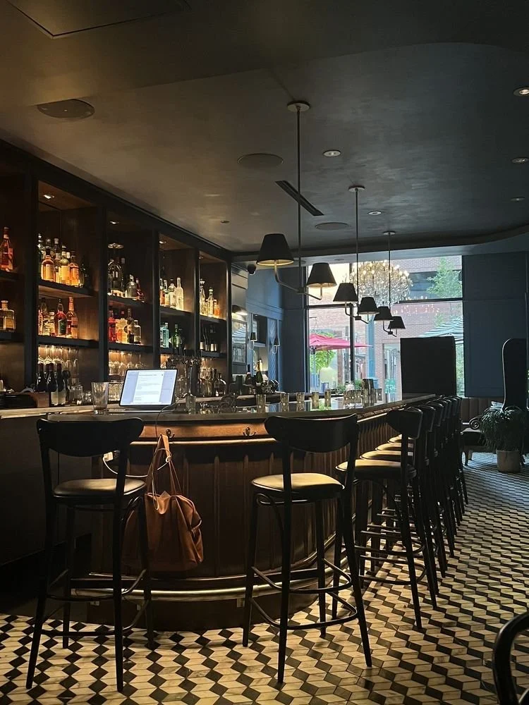 A dimly lit modern bar with a curved counter, high chairs, and shelves stocked with liquor bottles. A laptop and brown bag are placed on the bar, with geometric patterned flooring and pendant lights overhead. Large windows at the back show outdoor seating and greenery.